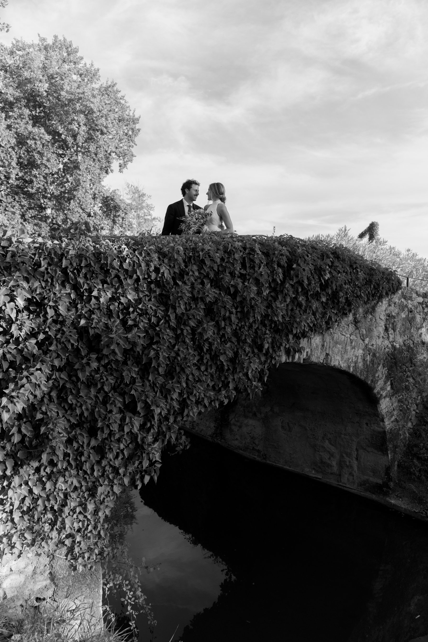 A black and white photo of a man and woman in wedding attire standing on a bridge over a body of water, gazing at each other.