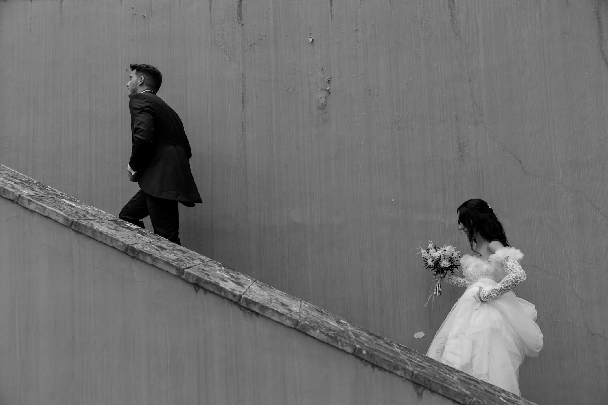 A black and white photograph of a bride in a wedding dress holding a bouquet walking behind a groom in a suit on a staircase.