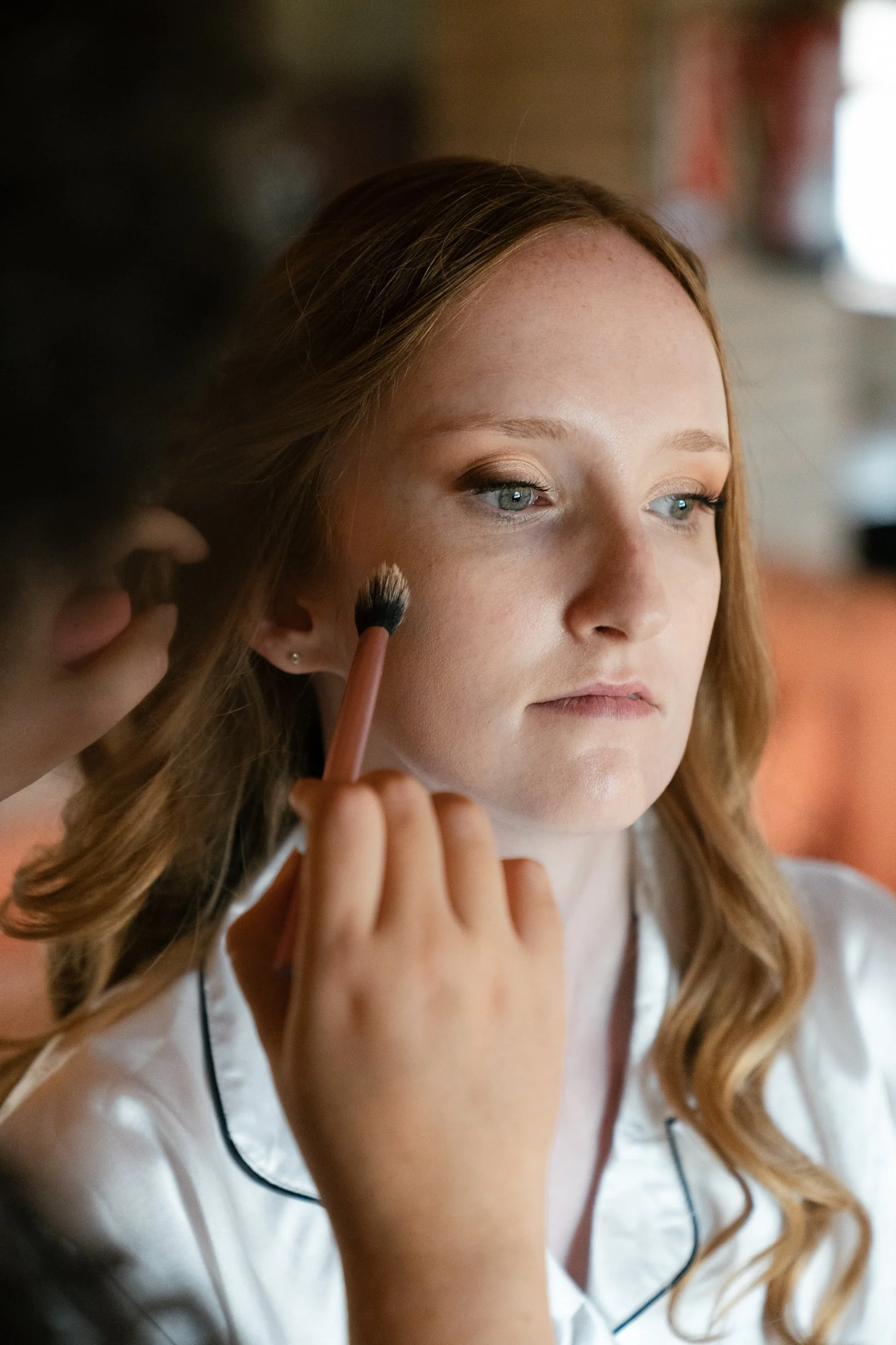 A woman with red hair getting her makeup done, with a makeup brush being applied to her face.