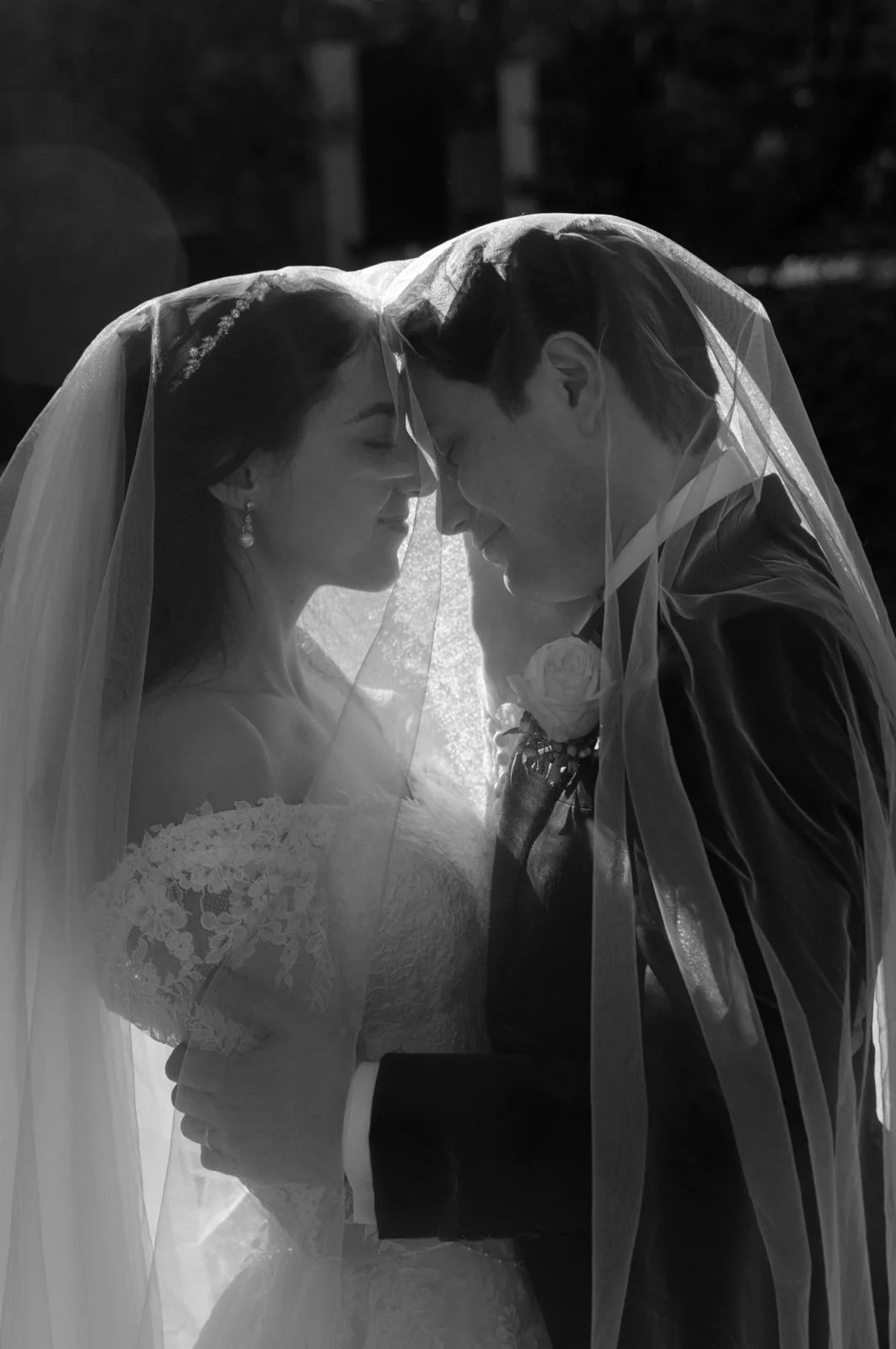 A black and white close-up photo of a bride and groom with their foreheads touching, smiling, under a veil during a wedding celebration.