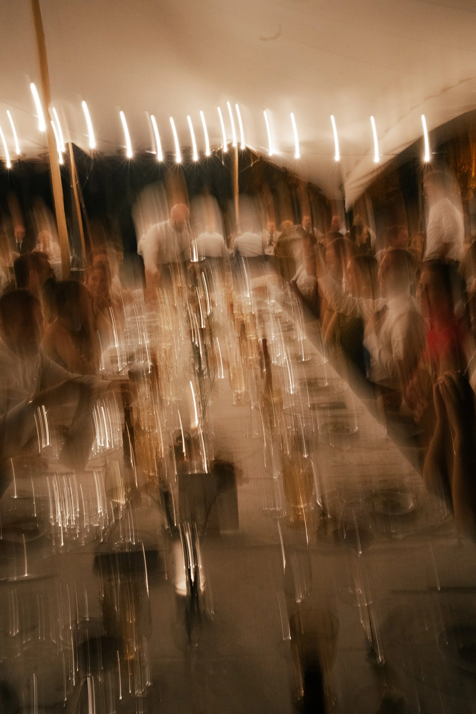 A blurred photo of a crowded indoor dinner event with a long table, glasses, and people socializing under warm lighting.