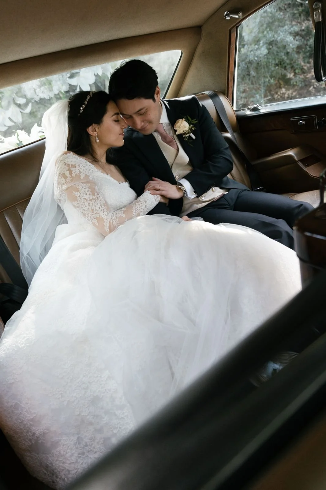 A bride and groom sitting close together in the backseat of a car, with their foreheads touching and eyes closed, appearing peaceful and emotional, during their wedding day.