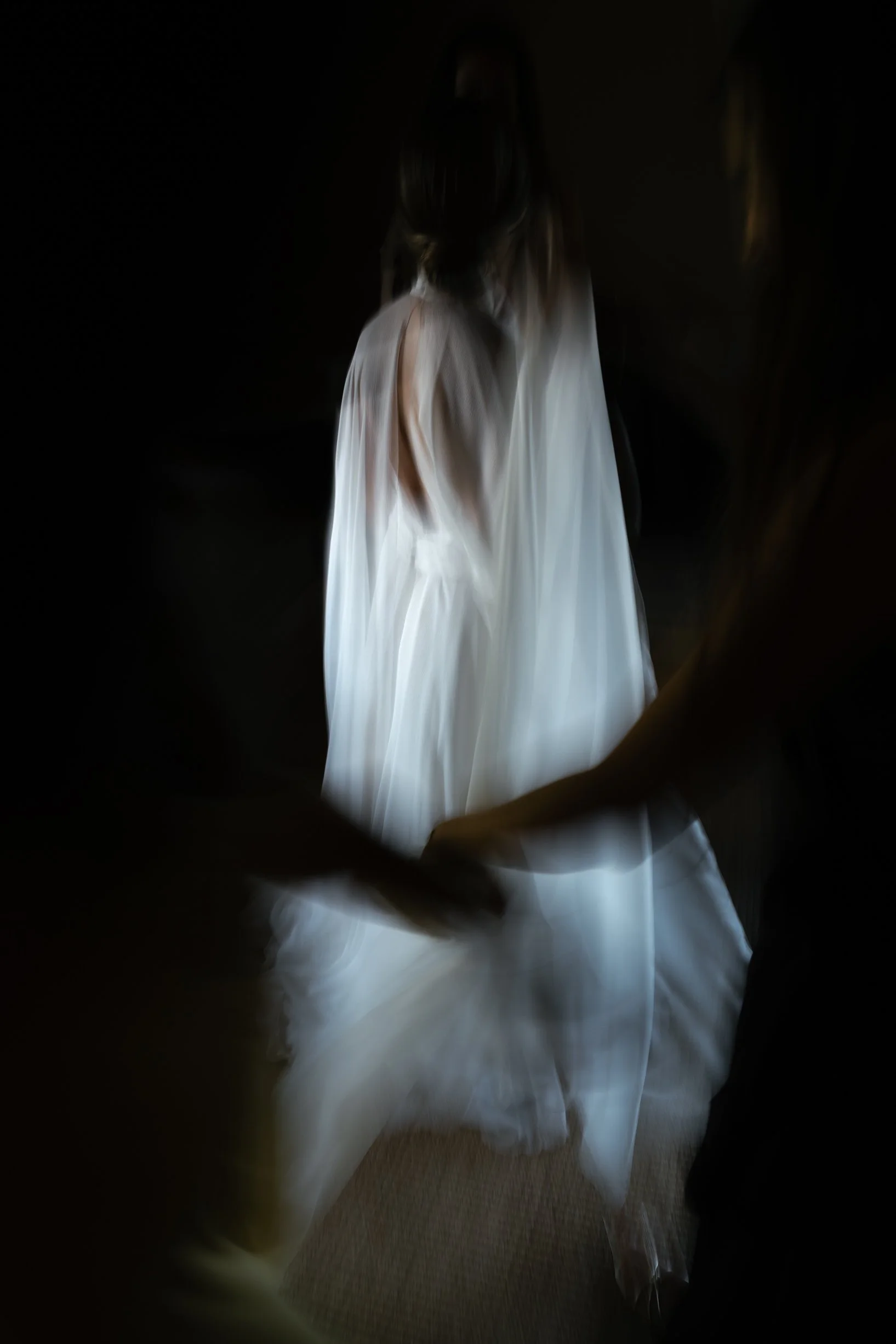 A woman in a white dress is being assisted by two people, holding her hand as she walks. The scene is dark with focused lighting on her dress.