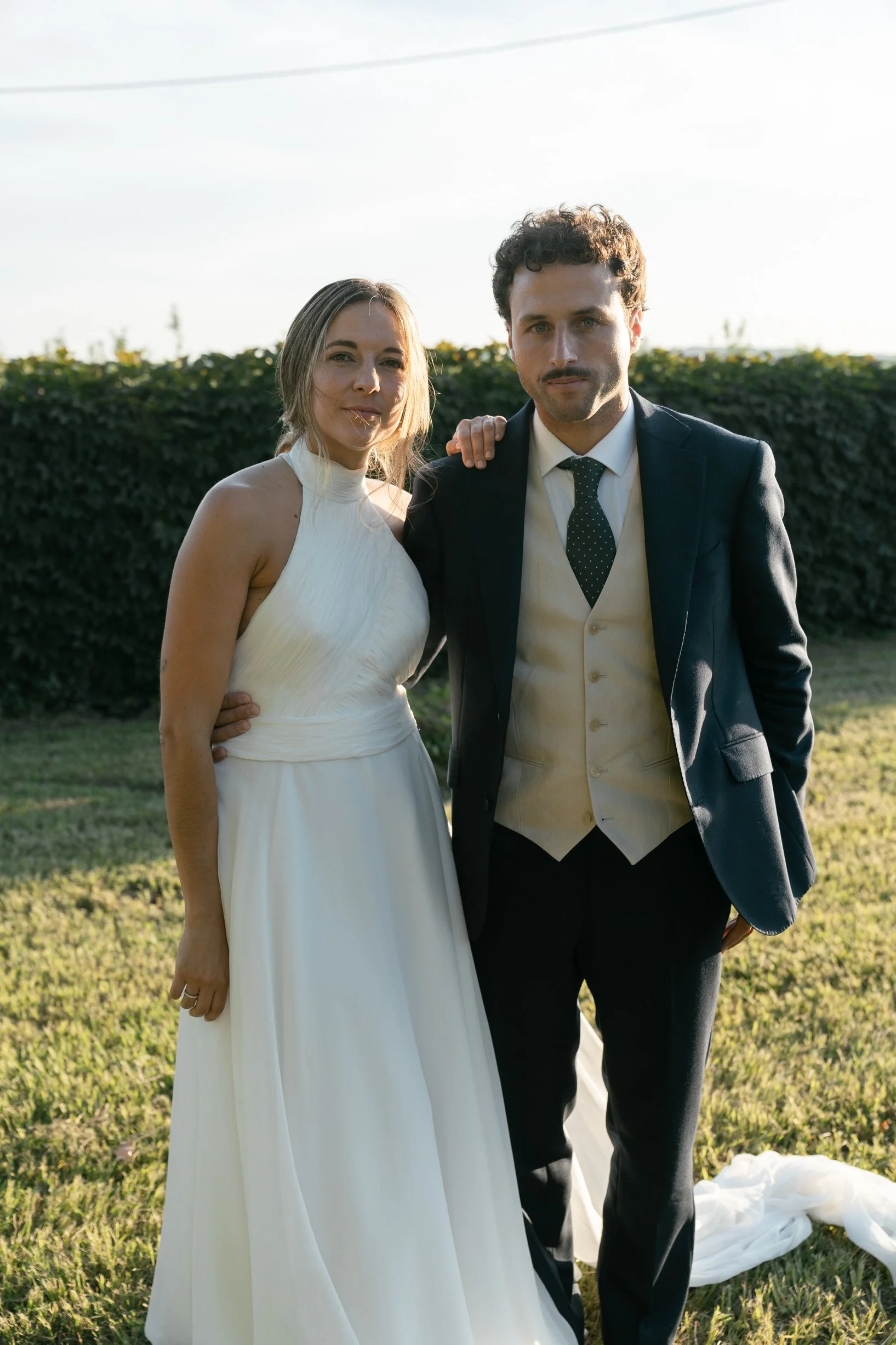 A bride and groom standing outdoors on grass, dressed in wedding attire, with the bride in a white gown and the groom in a black suit and vest. The bride has her arm around the groom's shoulder; they are looking at the camera with the sun setting beh