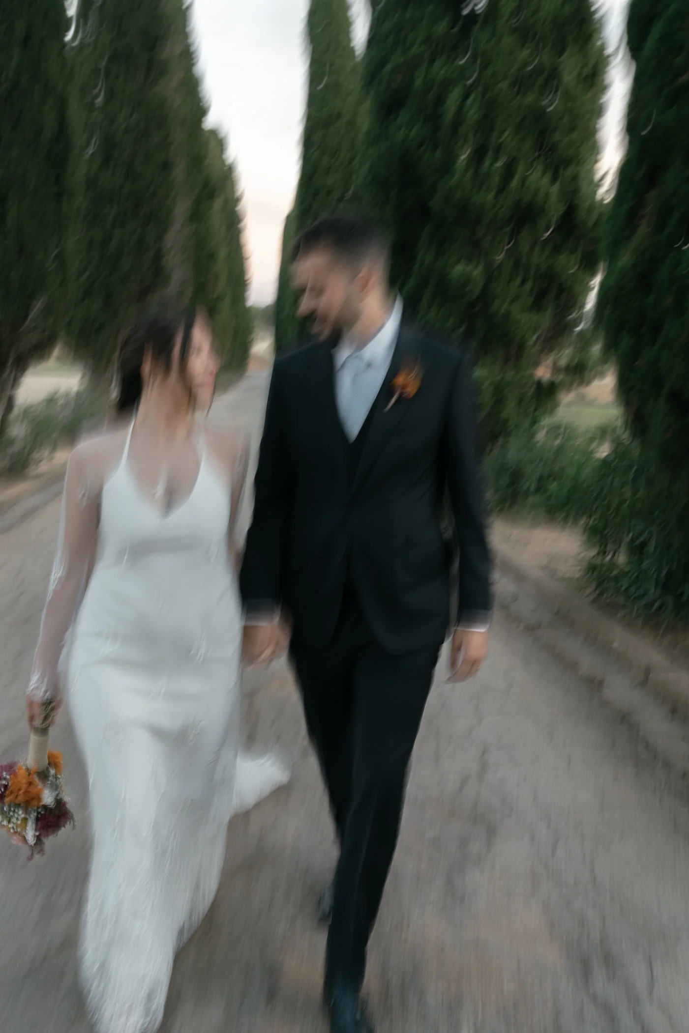A couple holding hands, walking outdoors with tall greenery on both sides, dressed in wedding attire, the woman in a white gown holding a bouquet, and the man in a black suit.