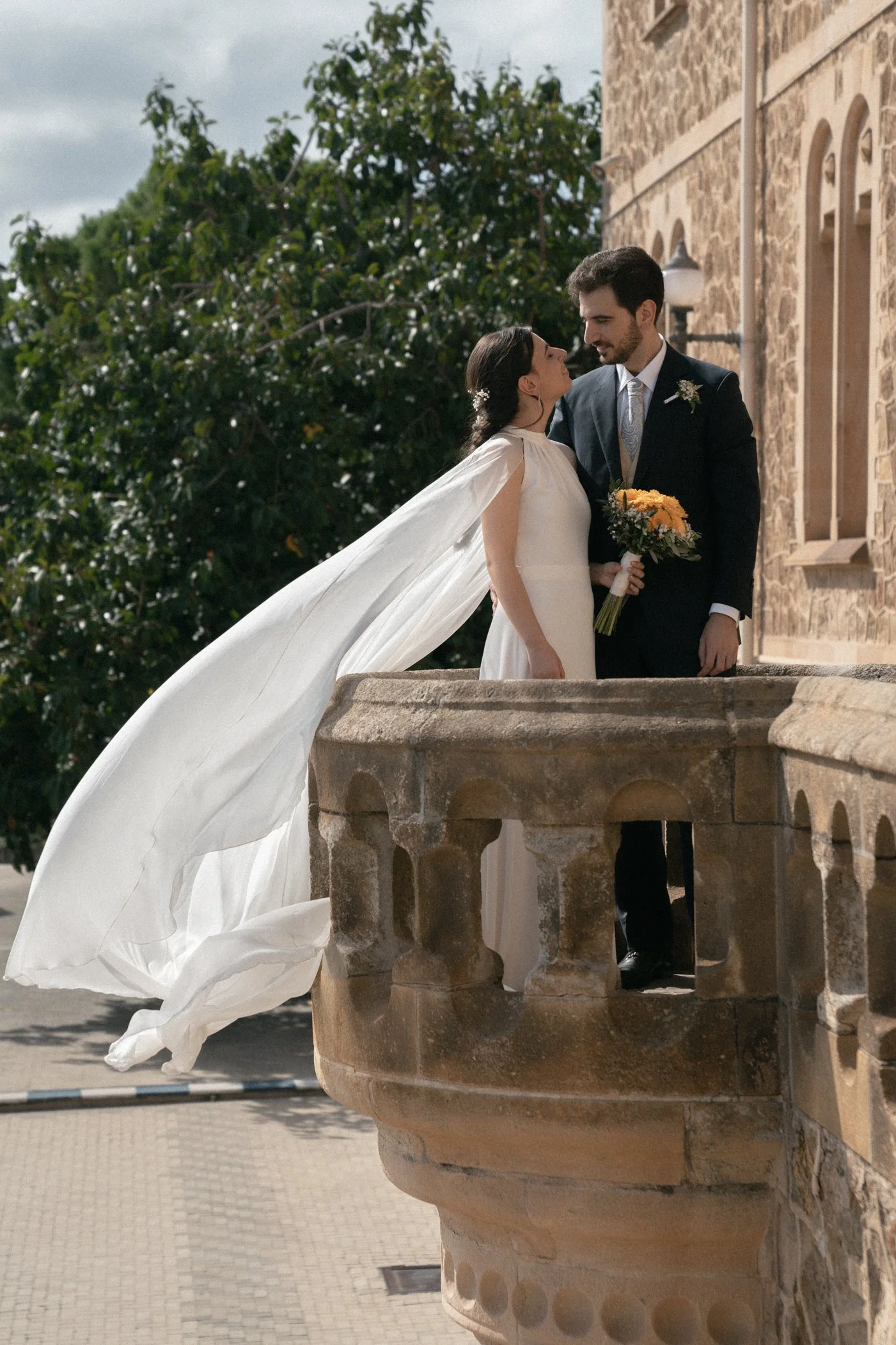 A bride and groom standing on a balcony in a wedding ceremony, with the bride's flowing white veil and holding a bouquet of yellow flowers, near a stone building and greenery in the background.