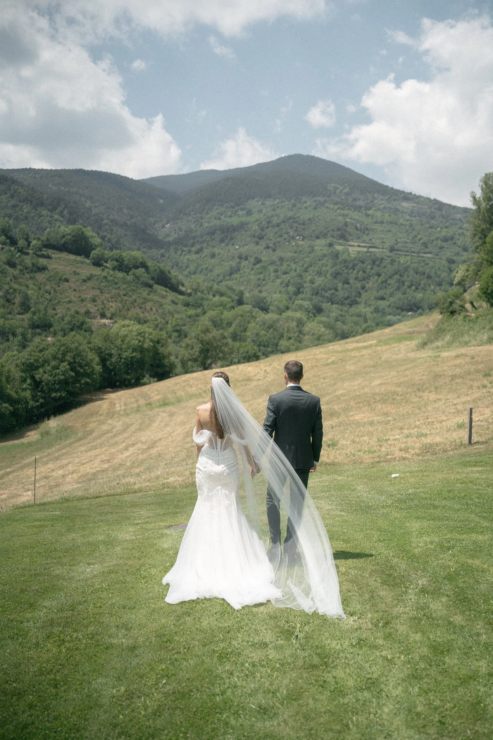 A bride and groom walking hand in hand across a grassy field with mountains and a cloudy sky in the background.