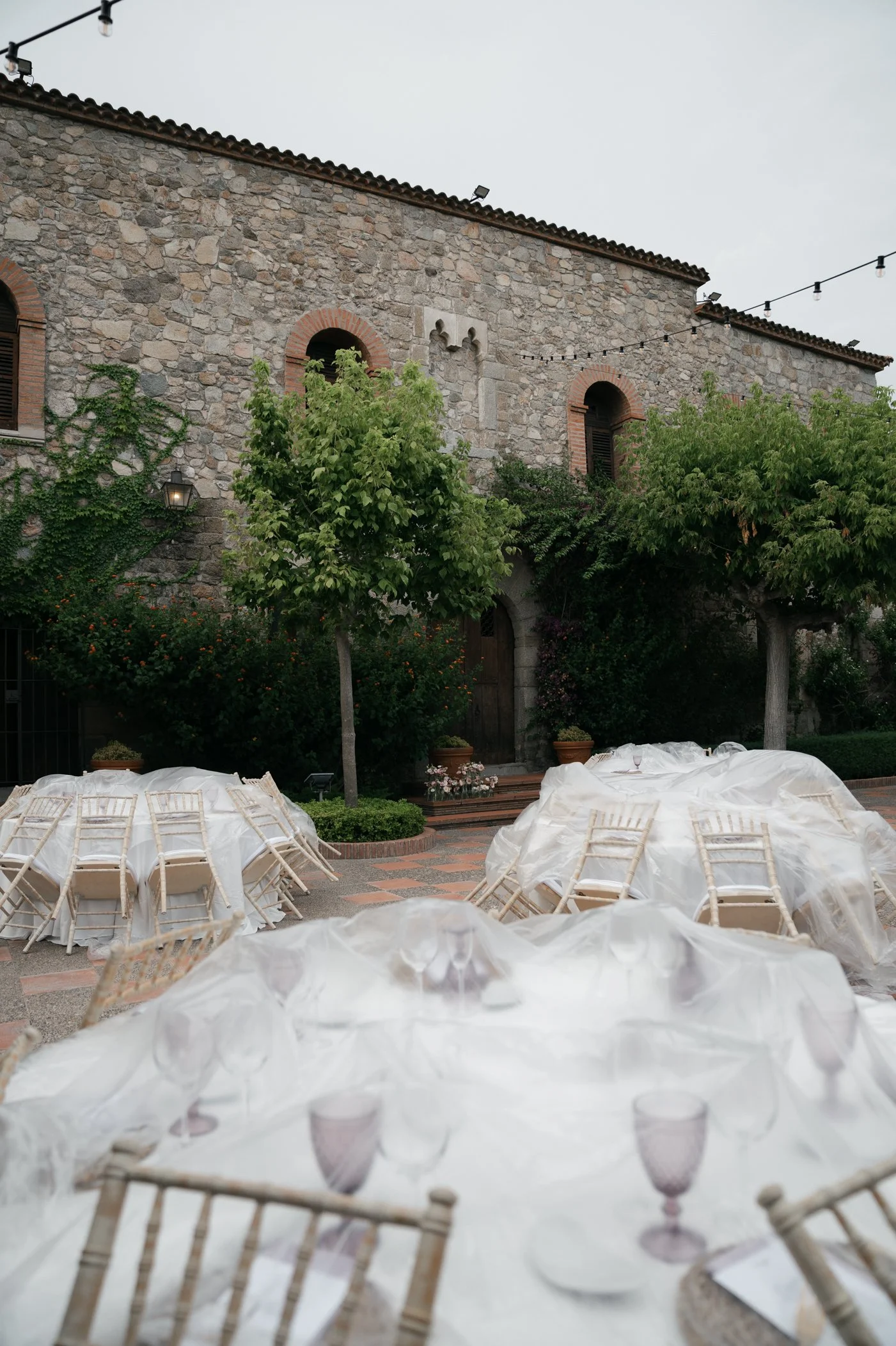 Outdoor event setup with tables covered in white tablecloths, surrounded by cream-colored chairs, in a courtyard with stone building, greenery, and string lights overhead.