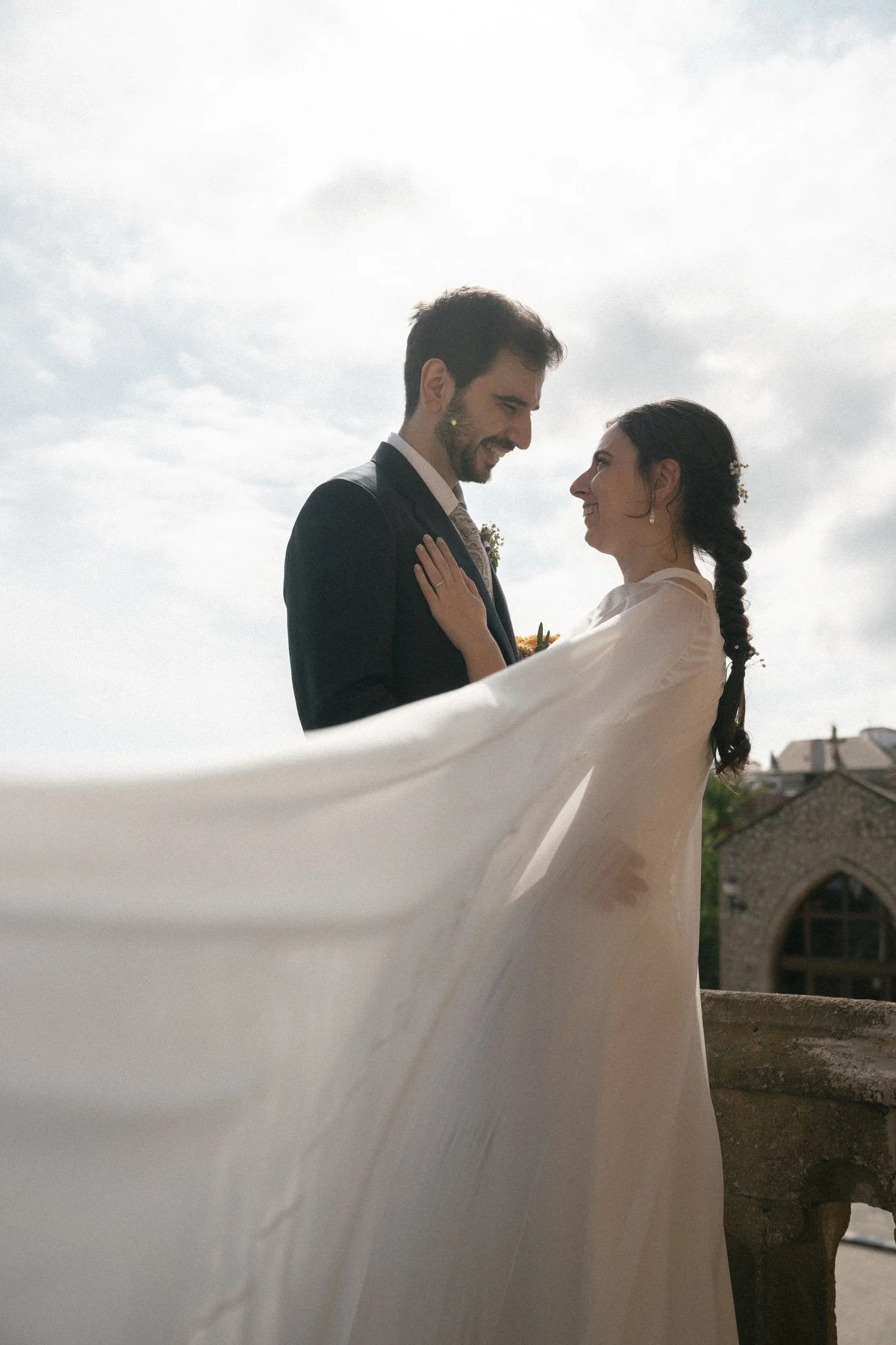 A bride and groom gazing at each other outdoors, with the bride's veil flowing in the wind and a historic building in the background.