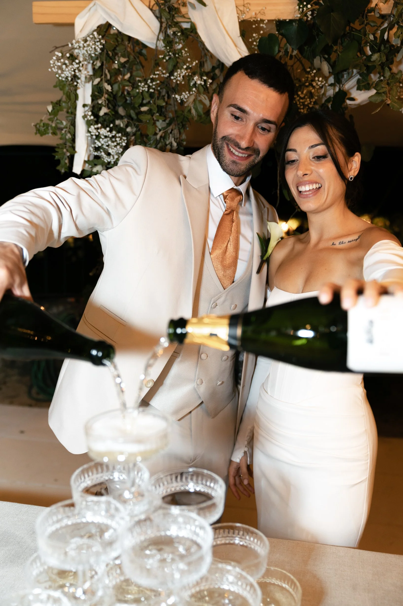 A newlywed couple pours champagne into a glass tower at their wedding reception, smiling and dressed in elegant wedding attire, with a floral and greenery backdrop.