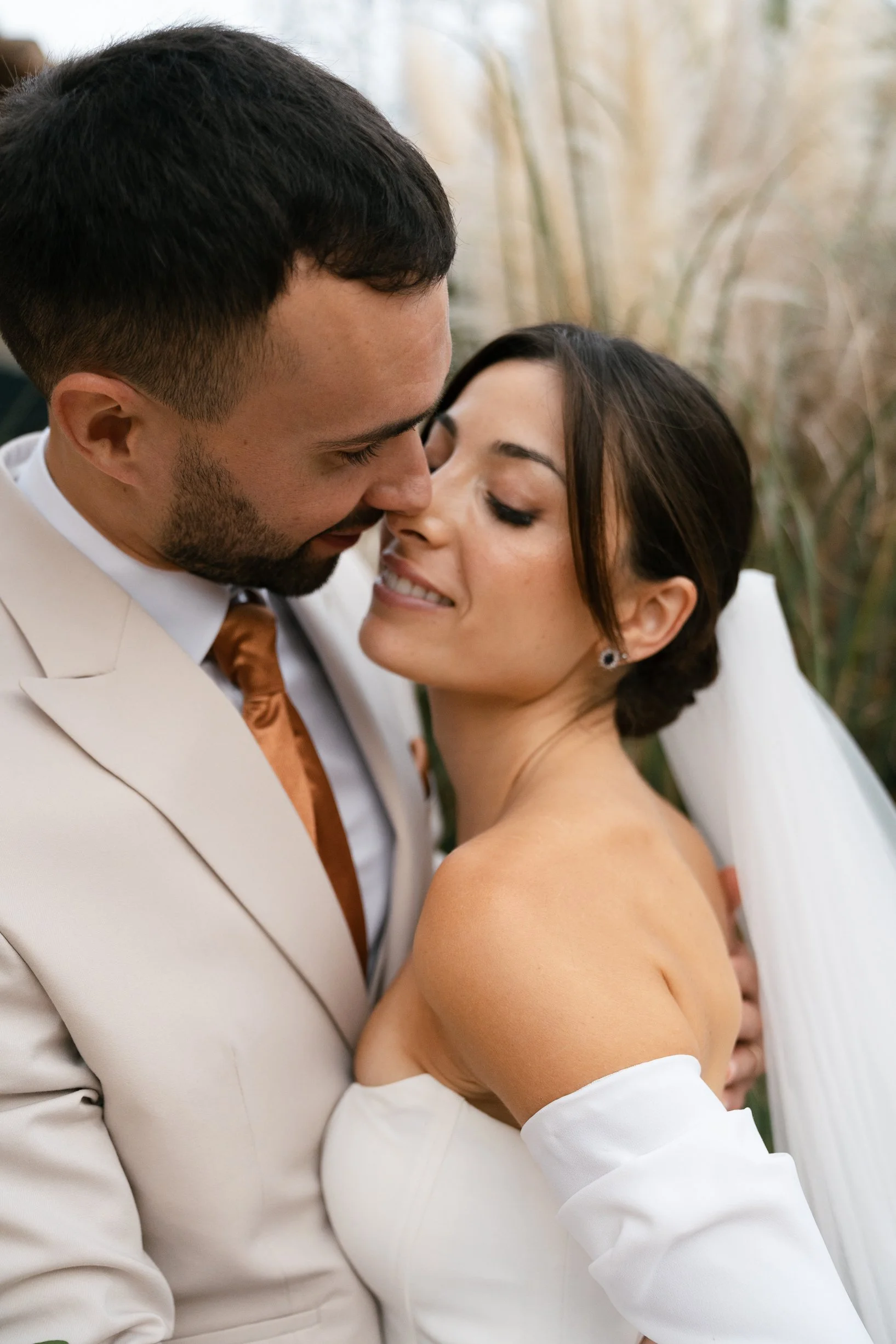 A couple, dressed in wedding attire, intimacy close-up outdoors, with tall grasses in the background.