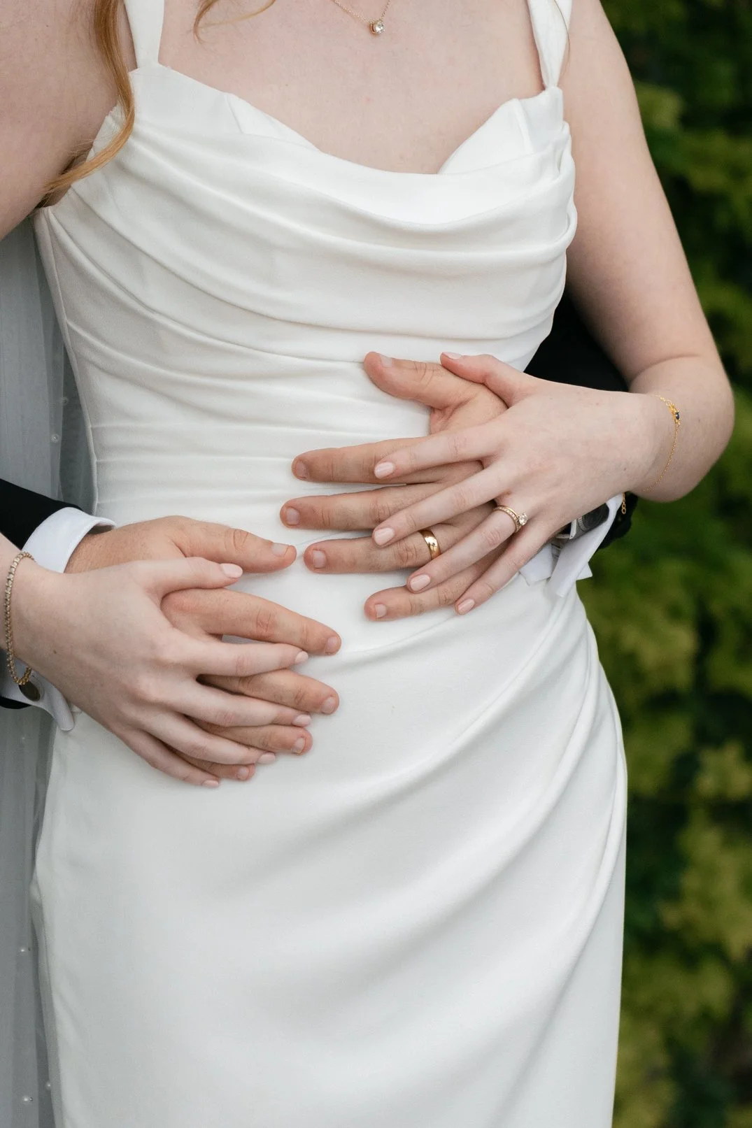 Close-up of a newlywed couple's hands resting on each other's waist, showing their wedding bands, with a bride in a white dress and some greenery in the background.
