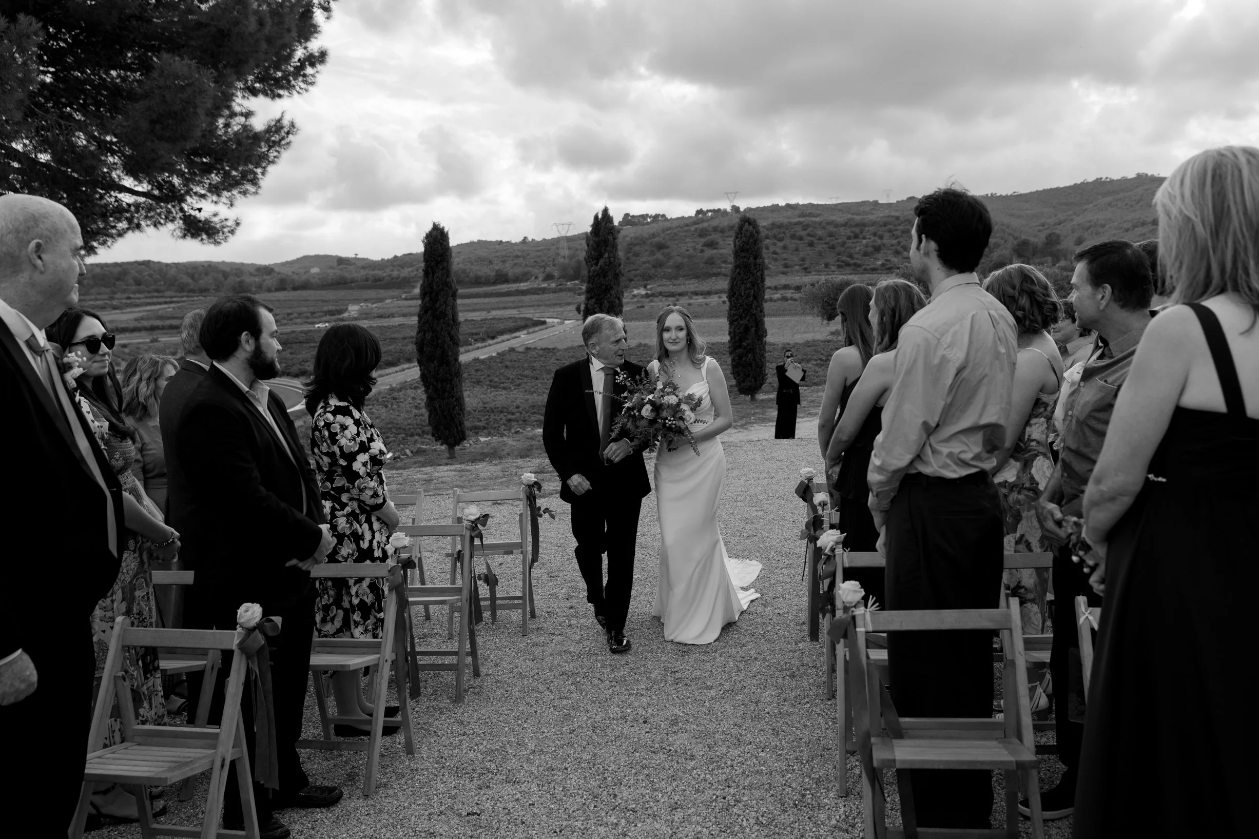 A black and white photo of a bride walking down the aisle with her father at an outdoor wedding ceremony, surrounded by guests on either side, with a scenic landscape and mountains in the background.