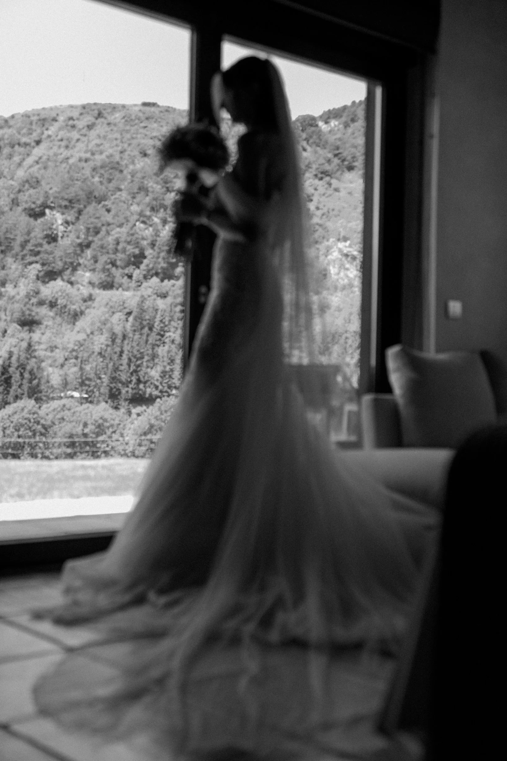 A woman in a wedding dress holding a bouquet, standing by a large window with a mountain landscape outside.