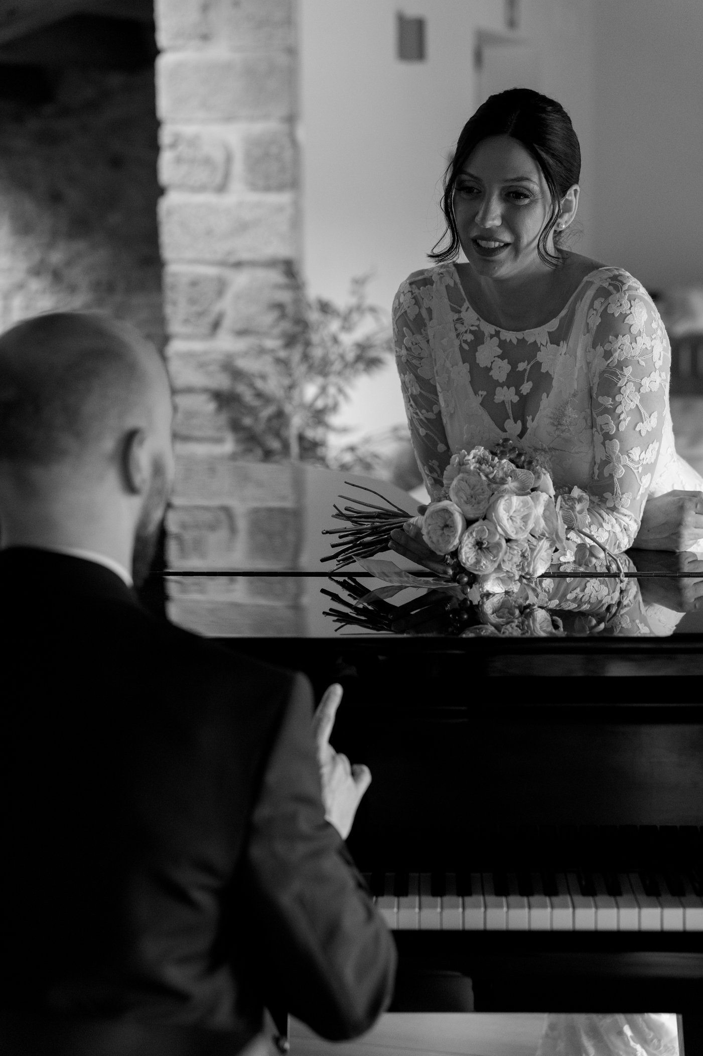 A woman in a lace dress holding a bouquet of flowers, sitting across from a man in a suit at a piano, in a cozy indoor setting.
