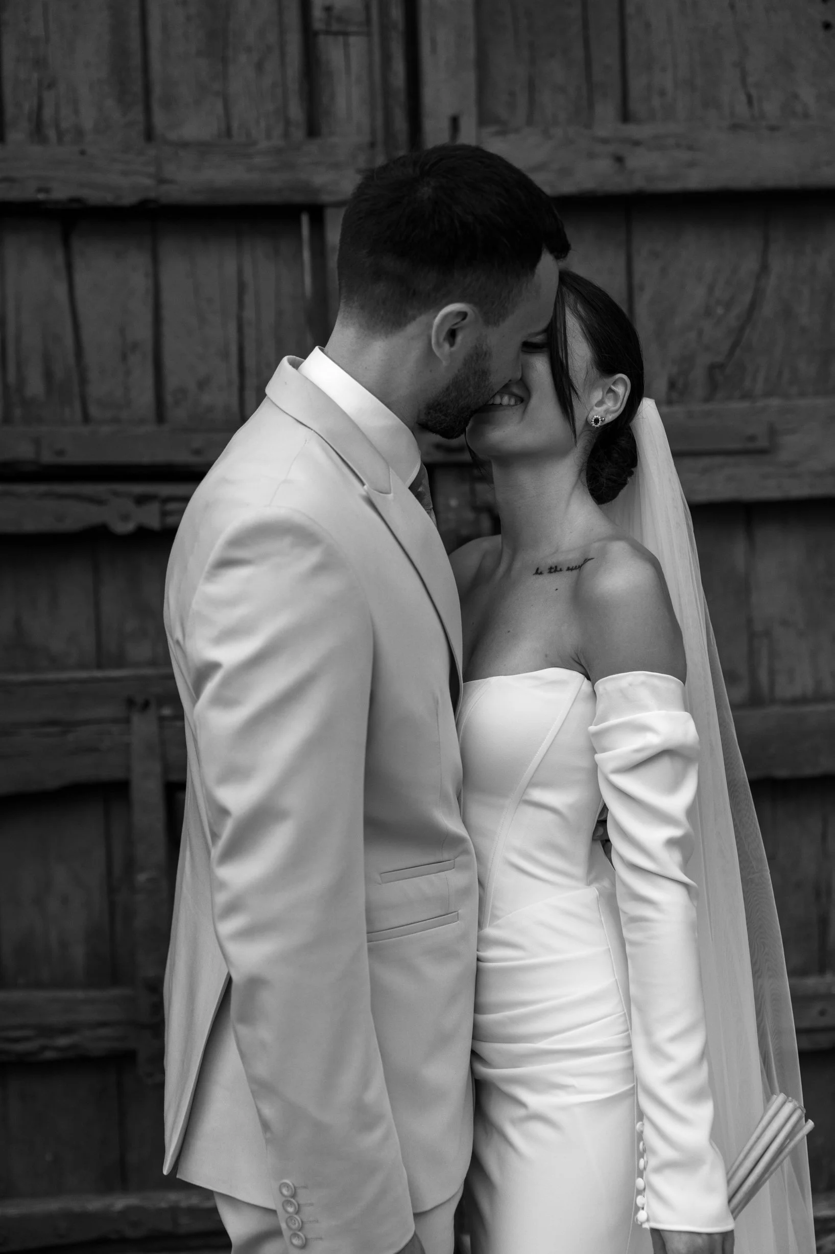 Black and white photo of a bride and groom close together, smiling, with their foreheads touching, in front of a wooden wall.