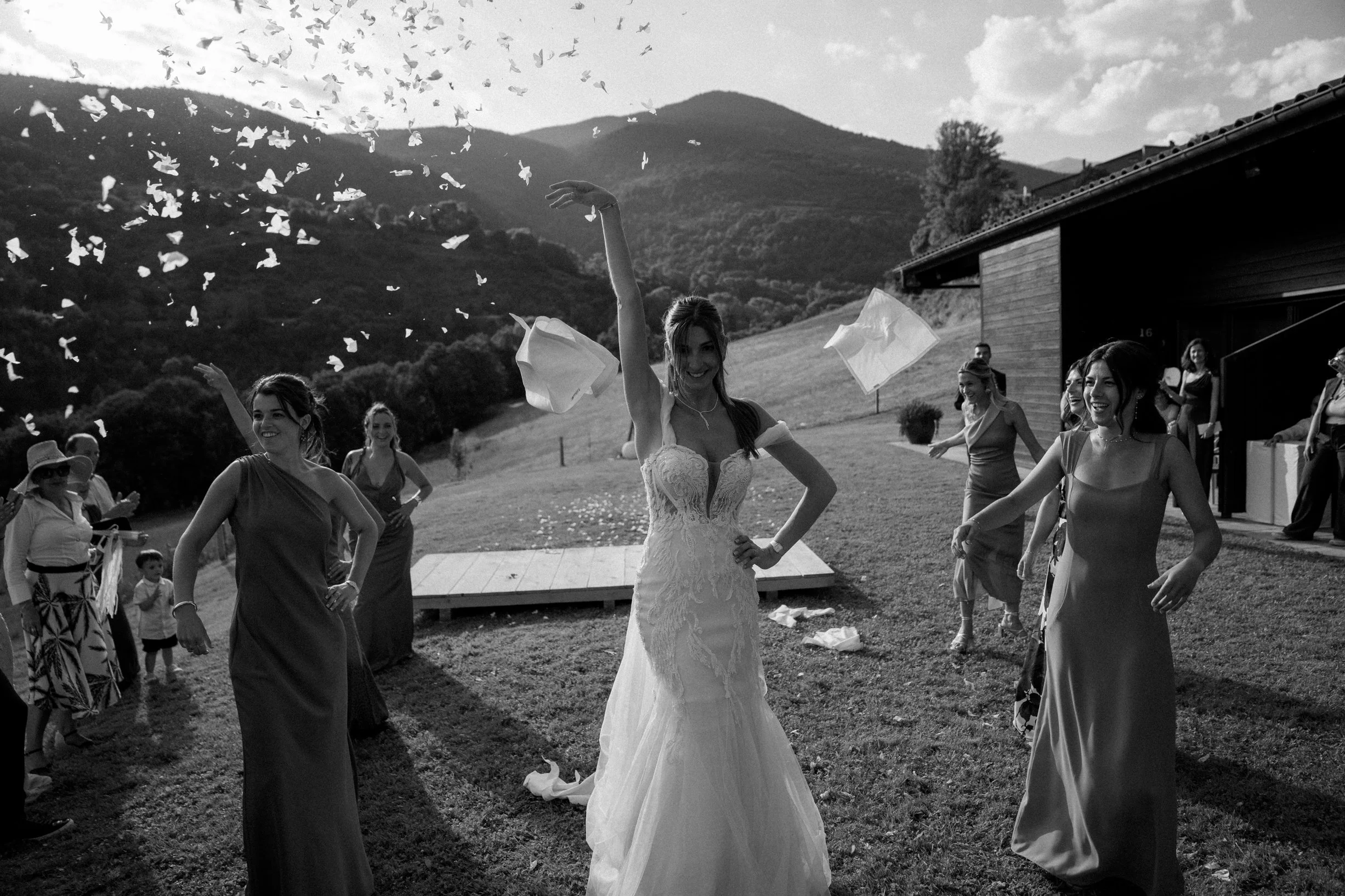 Bride and bridesmaids celebrating outdoors on a grassy hillside, throwing confetti, with mountains in the background.