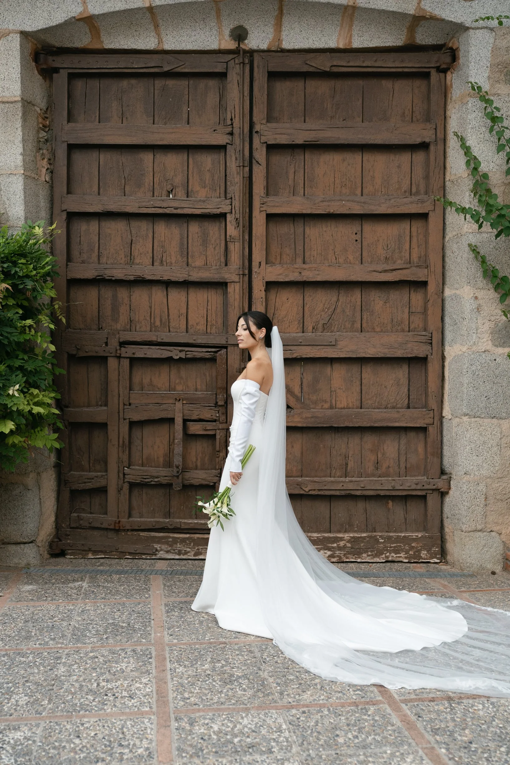 Bride standing in profile with a bouquet of calla lilies against a wooden gate with stone framing.