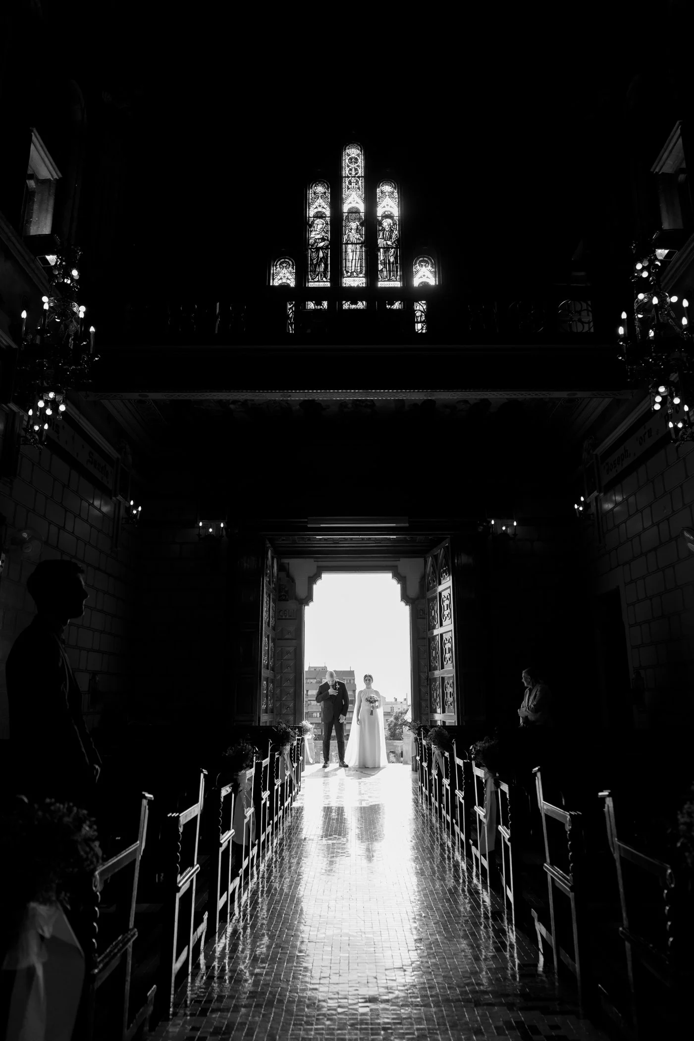 Silhouette of a bride and groom standing at the entrance of a church during a wedding ceremony, with sunlight shining from outside, stained glass windows, and guests seated along the aisle.