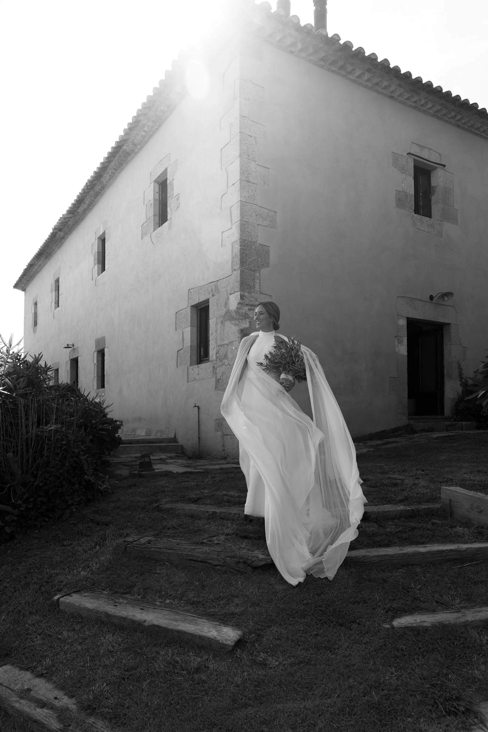 A woman in a white dress holding a bouquet of flowers, walking outside near a large stone building with small windows, captured in black and white.