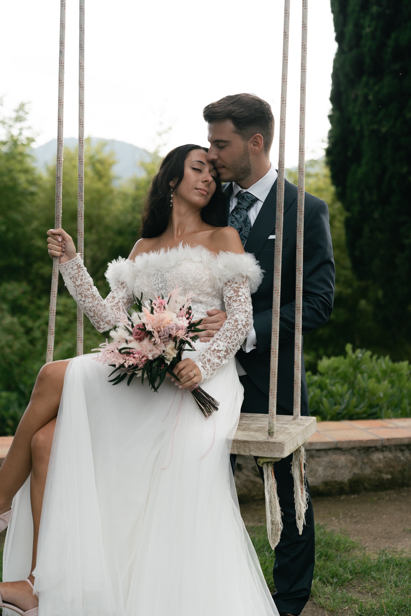 A bride and groom sitting on a wooden swing outdoors, with the bride holding a bouquet of pink and white flowers, dressed in a white wedding gown with lace sleeves, and the groom in a dark suit, in a green, natural background.