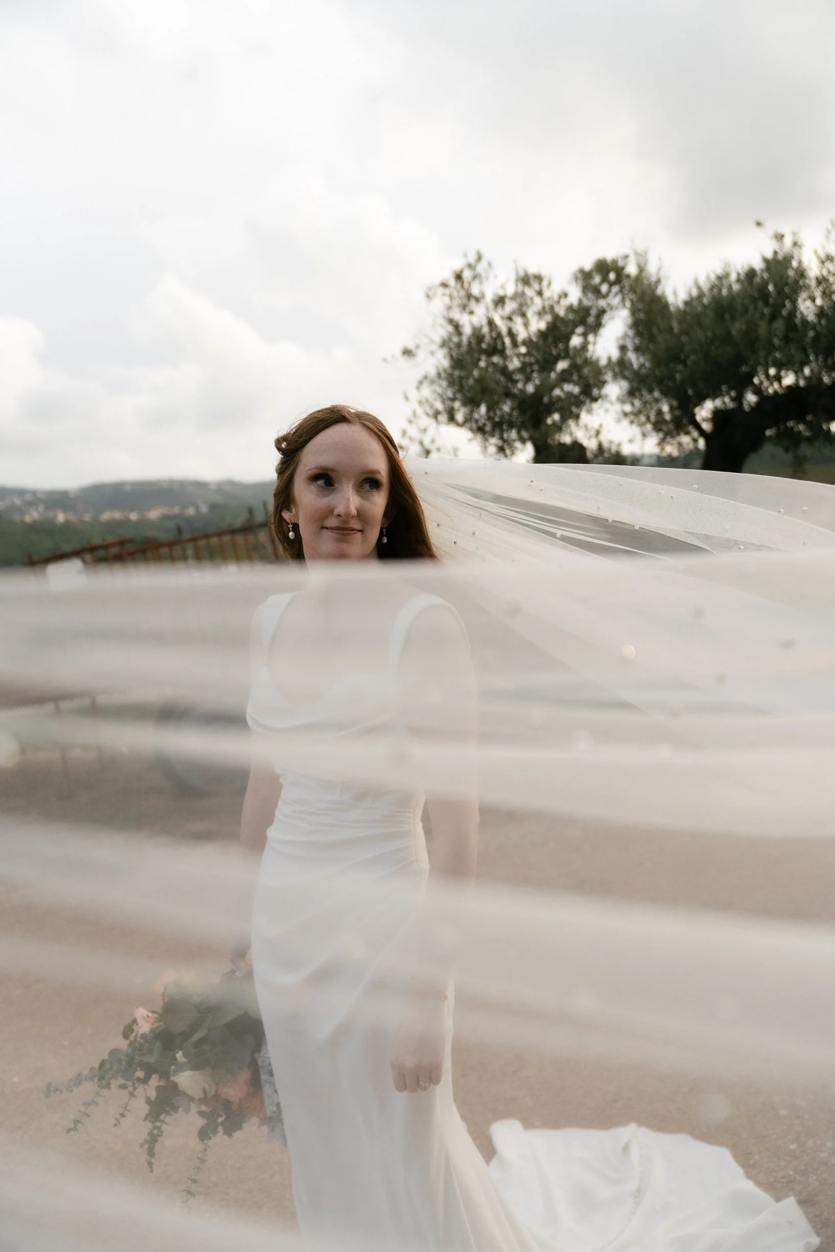A bride standing outdoors, holding a bouquet of flowers, with her veil flowing in the wind, trees and cloudy sky in the background.
