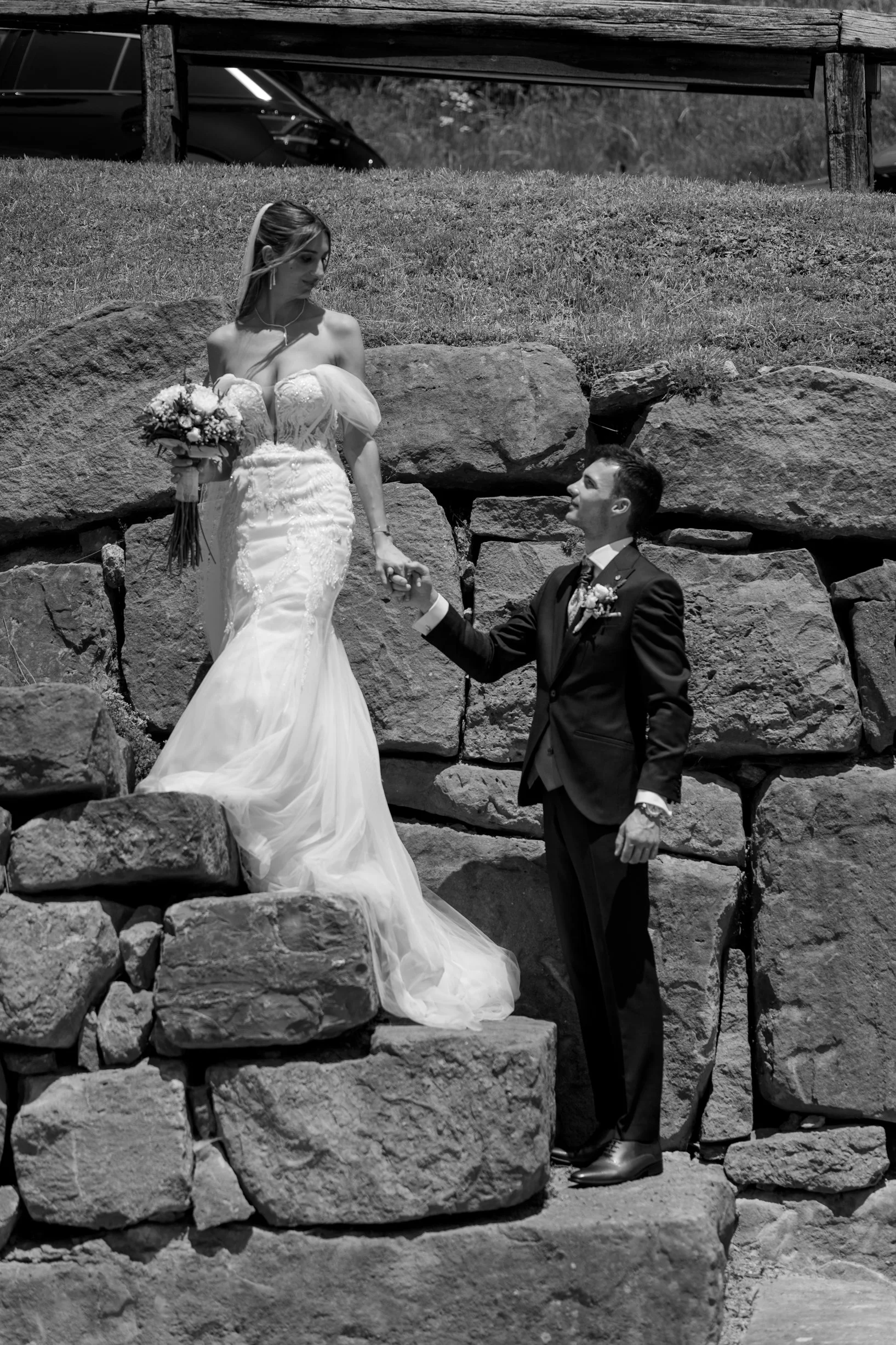 A black and white photo of a bride and groom outdoors, with the bride standing on rocks and the groom on the ground, holding her hand.