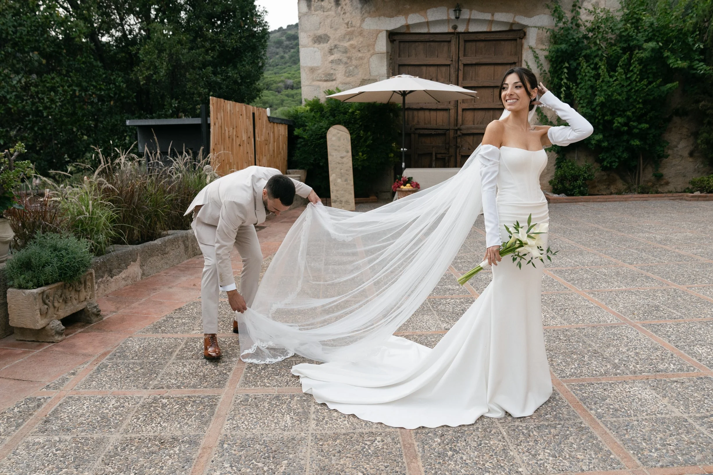 A bride in a white off-shoulder wedding gown holding a bouquet of white flowers with greenery, smiling as a man adjusts her long veil outdoors with a stone building, wooden door, and greenery in the background.