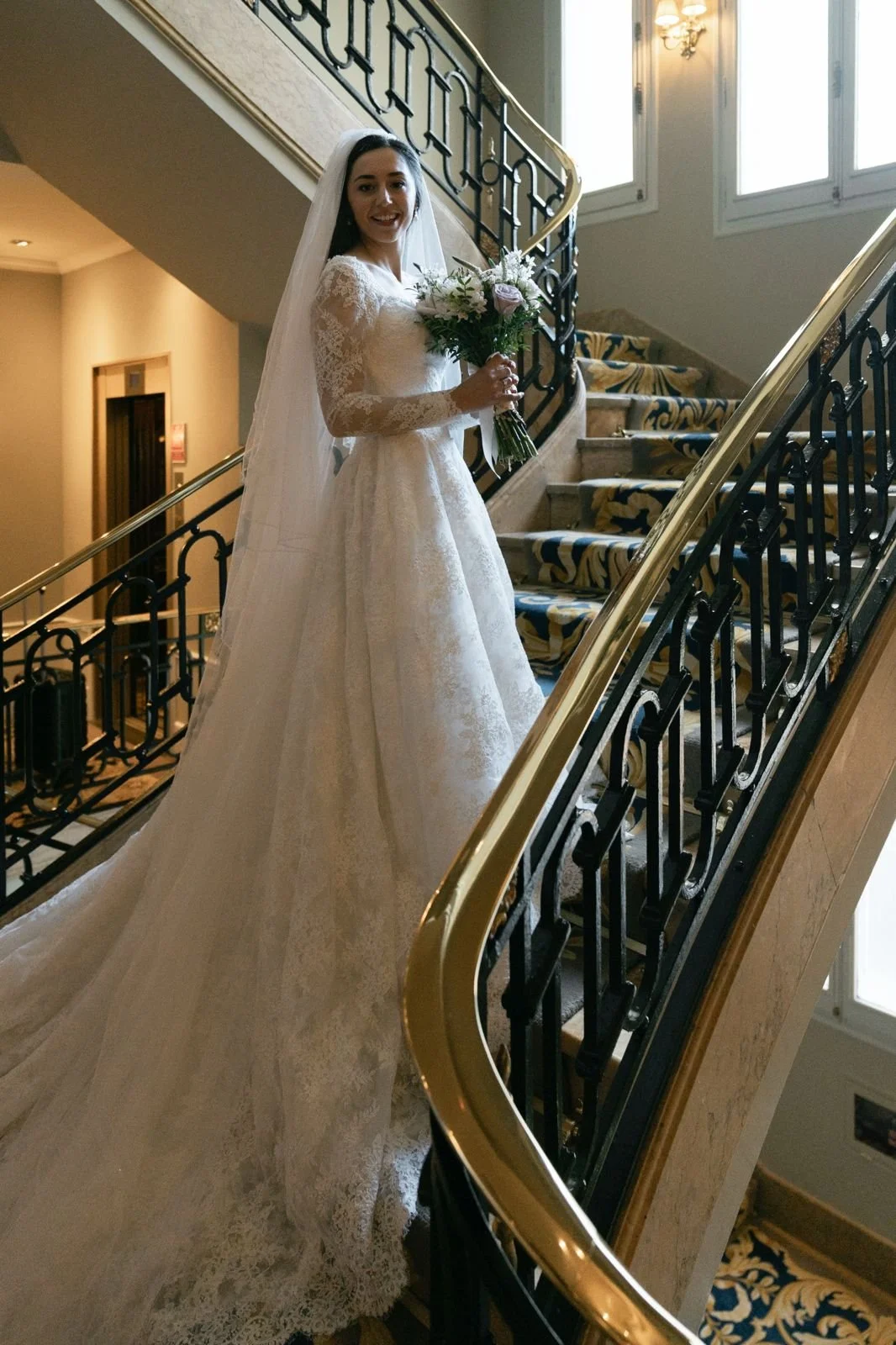 A bride in a white lace wedding dress holding a bouquet standing on a staircase in an elegant indoor setting.
