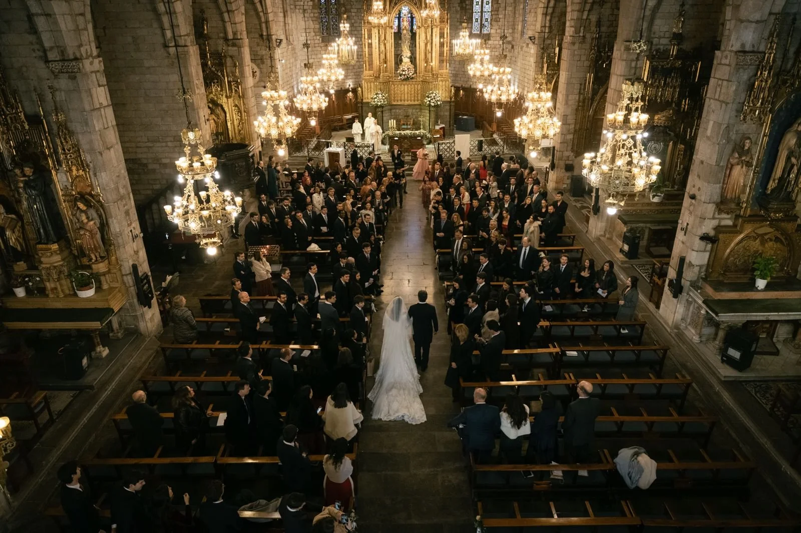 A wedding ceremony taking place in a grand cathedral with chandeliers, religious statues, and ornate decorations. The bride and groom walk down the aisle, surrounded by seated guests.