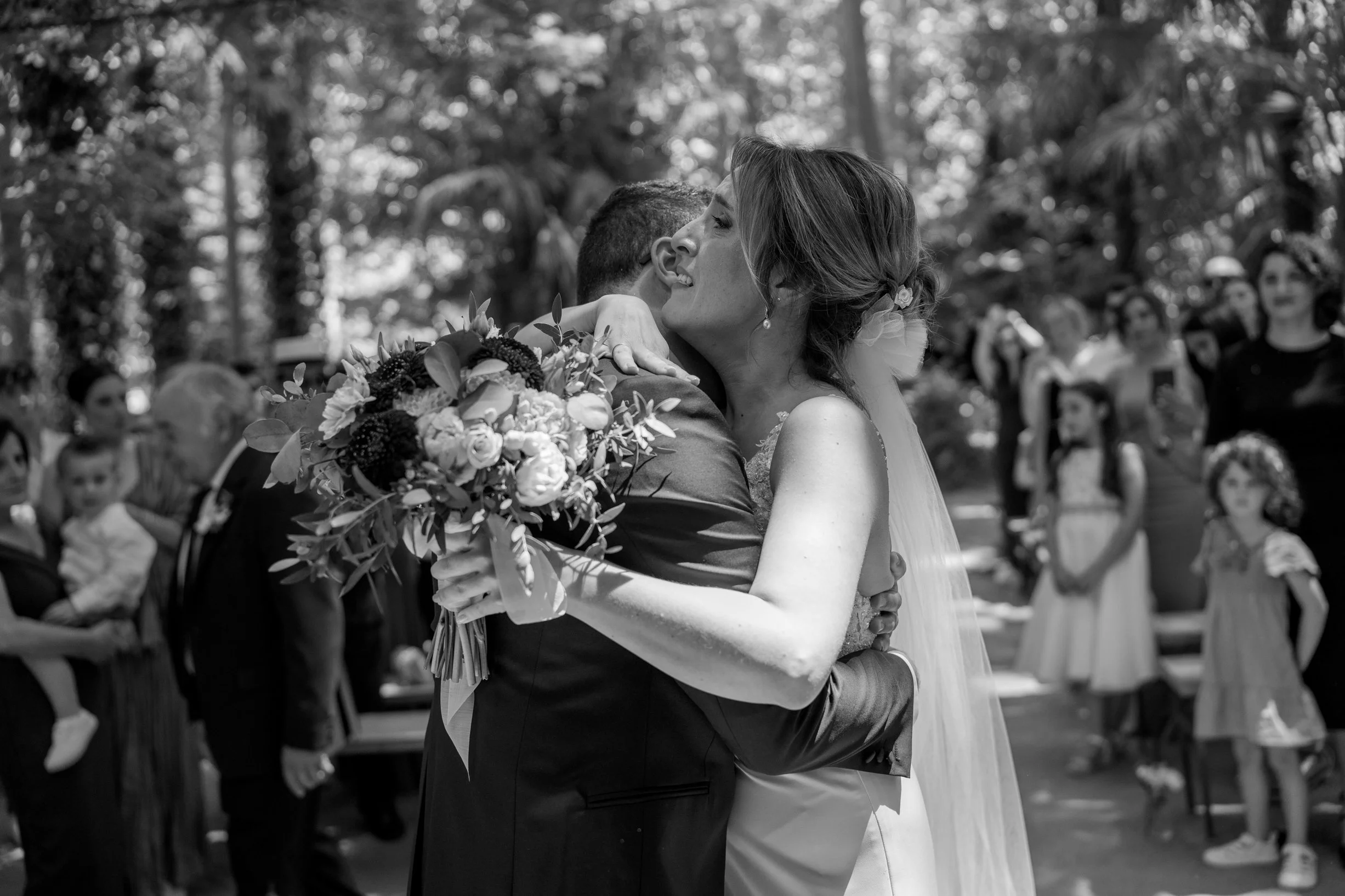 A bride and groom embrace during their wedding in an outdoor setting, with guests watching in the background.