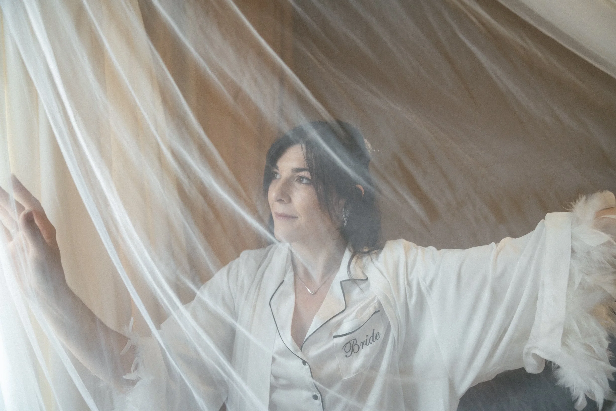 A bride in a white satin pajama top with black piping and embroidery that says "Bride" on the chest, standing behind a sheer veil with her arms outstretched, posing gracefully.