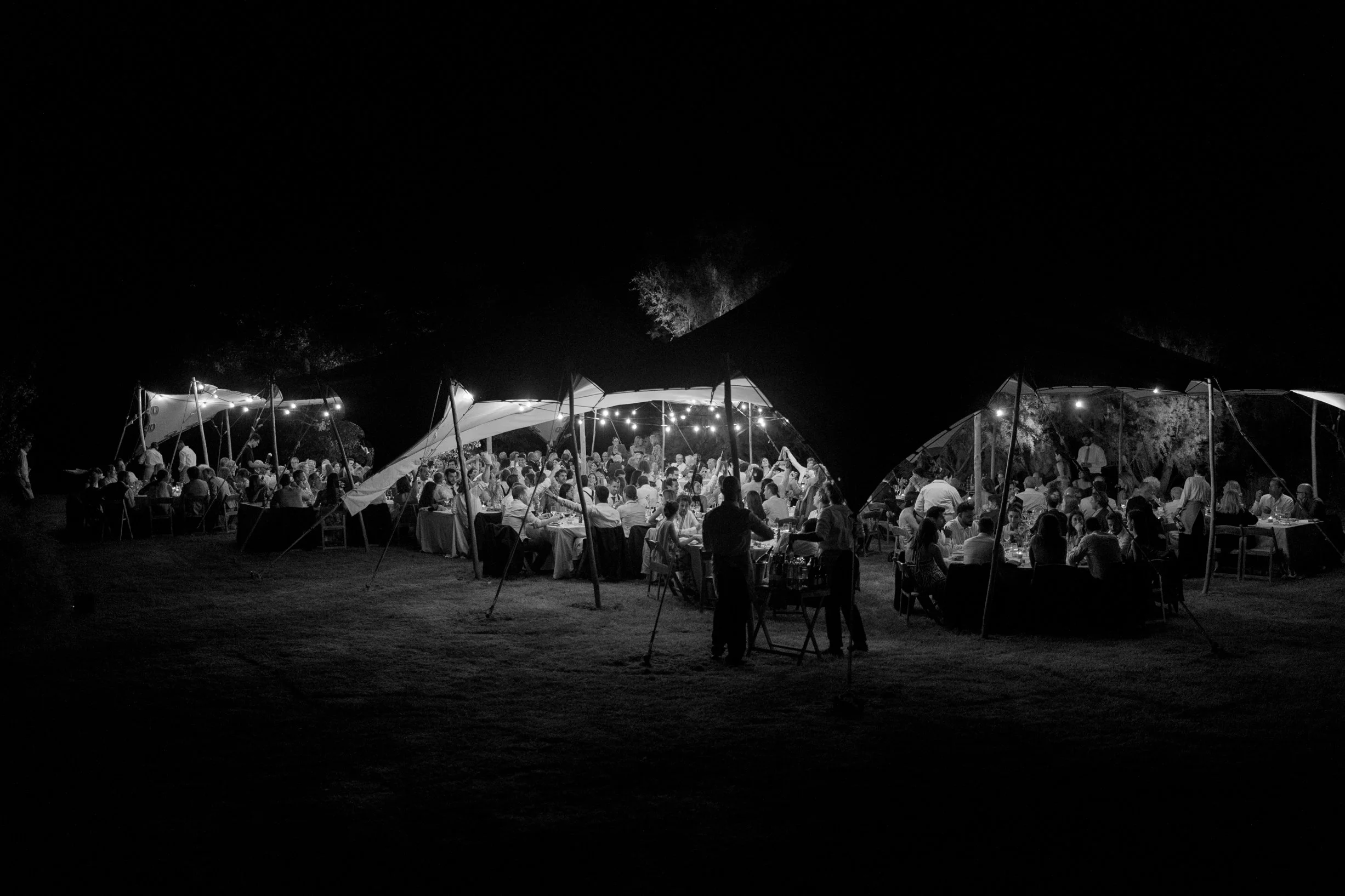 Nighttime outdoor event with people dining under tents with string lights, surrounded by darkness.