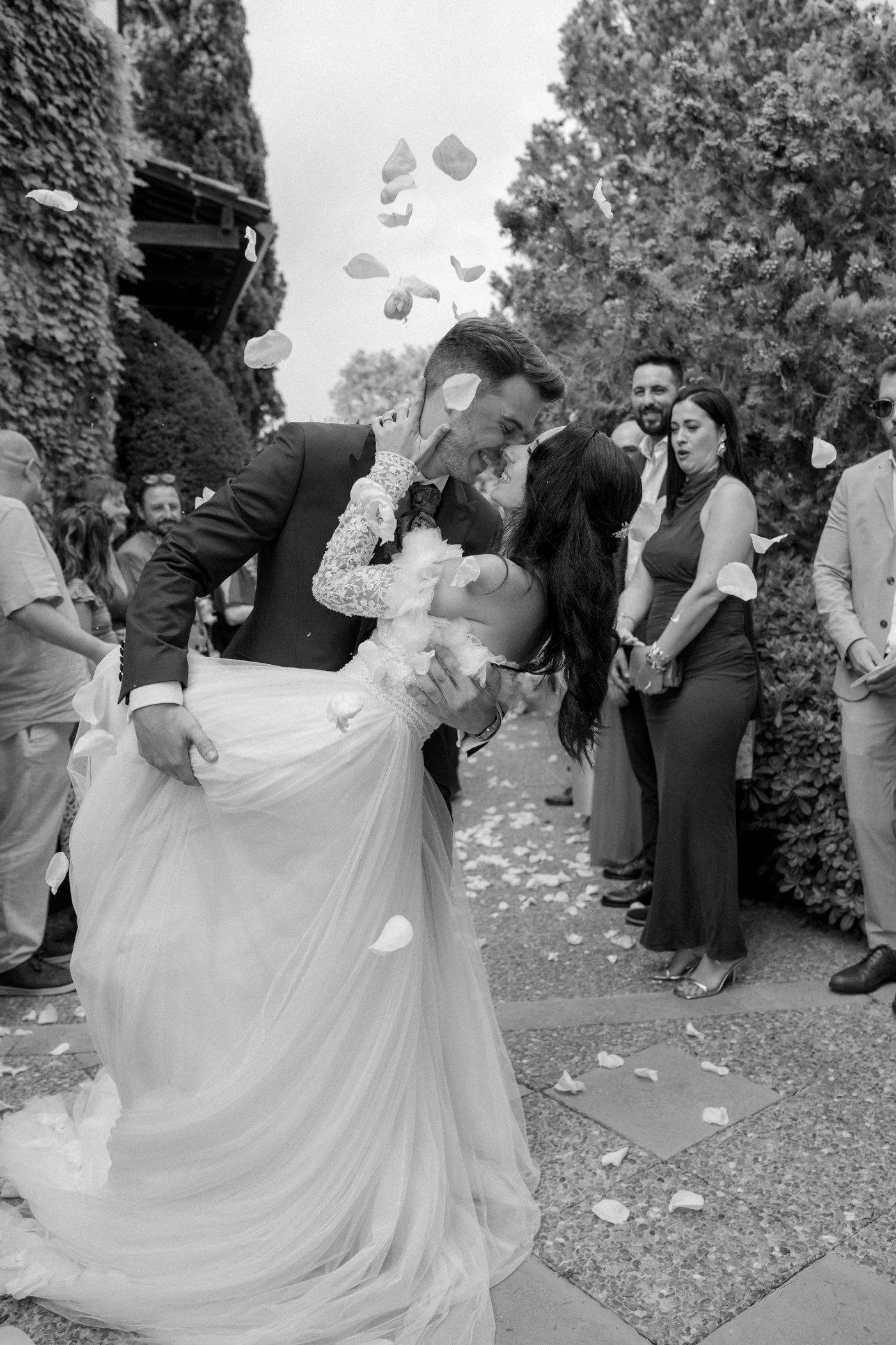A bride and groom sharing a kiss and dancing at their outdoor wedding, surrounded by friends and family, with flower petals falling around them.