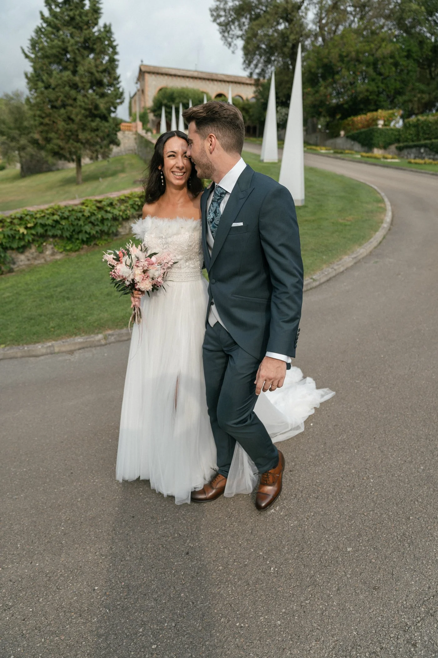 A bride and groom sharing a moment outdoors at their wedding, with greenery and modern sculpture art in the background.