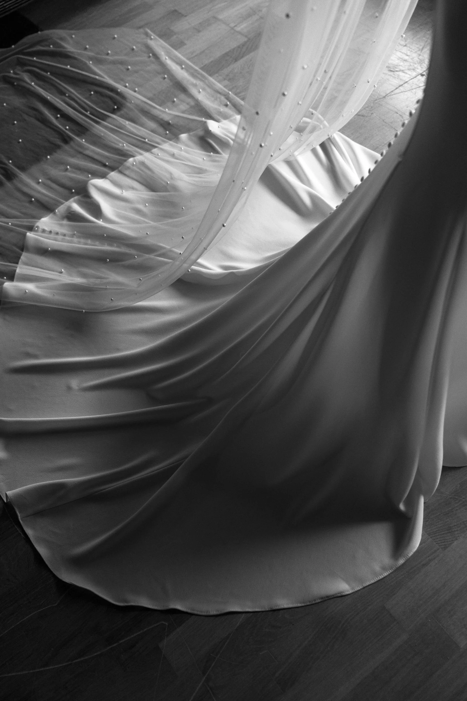 Close-up of a luxurious fabric draped on a wooden floor, possibly part of a dress or curtain, with delicate beaded embellishments and sheer overlay.