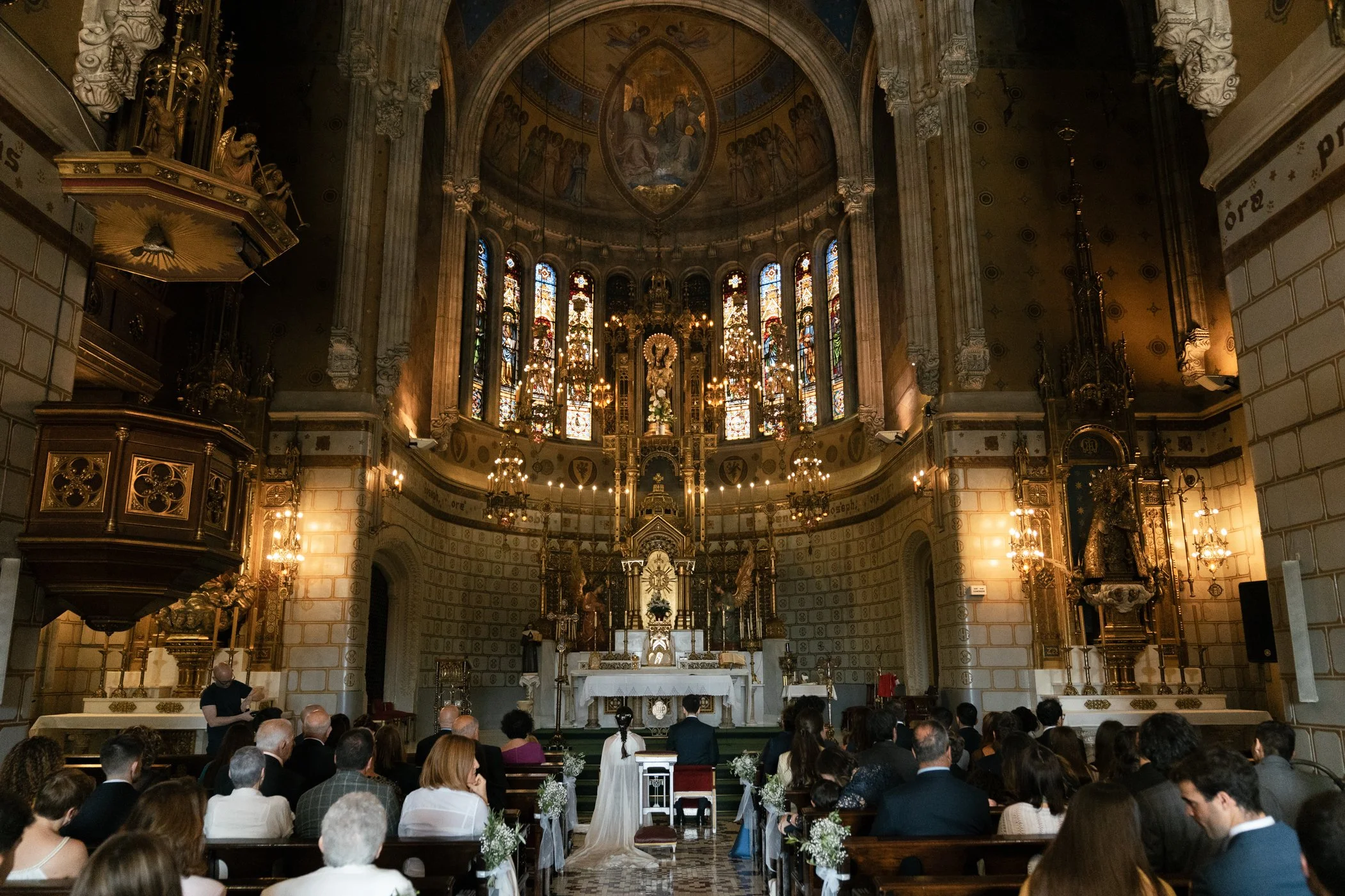 A wedding ceremony inside a decorated church with stained glass windows, an altar, and numerous guests seated in pews.