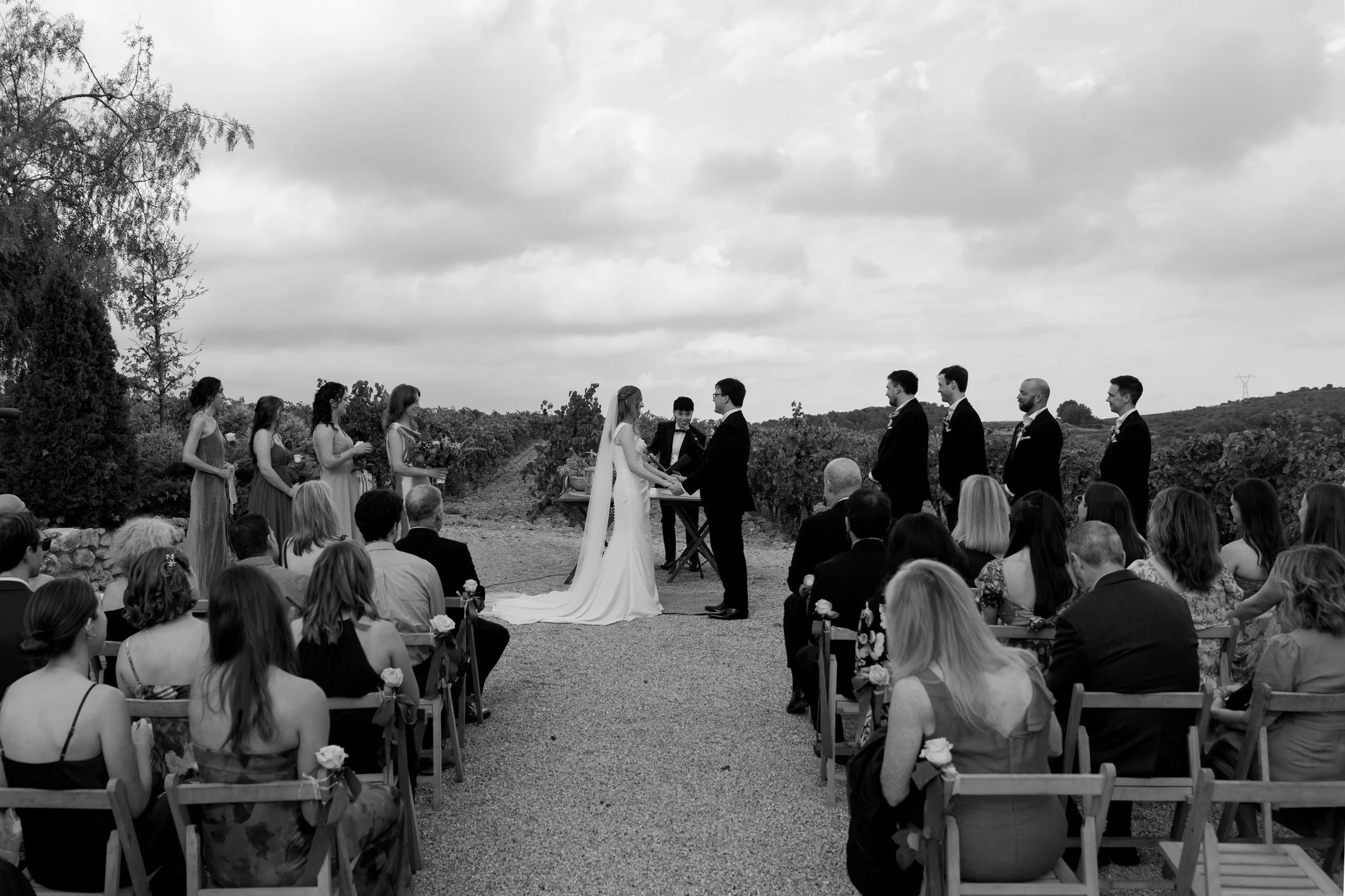 A black and white photo of an outdoor wedding ceremony with the bride and groom holding hands at the altar, surrounded by bridesmaids and groomsmen, with guests seated observing the event.