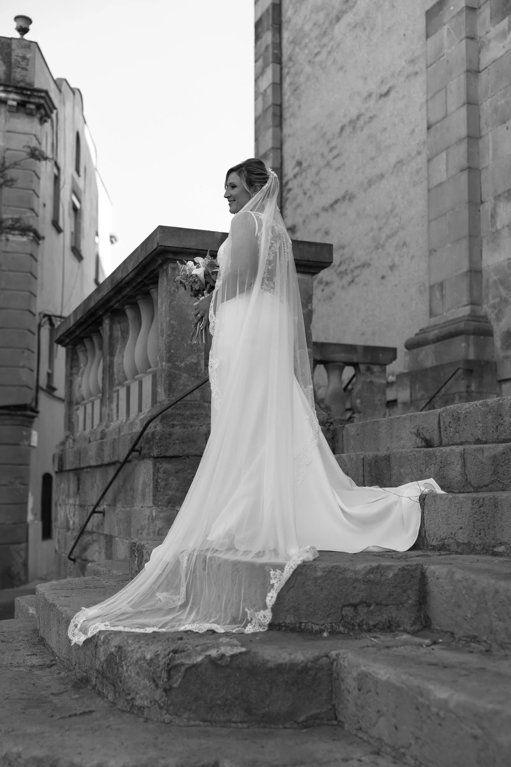A bride in a wedding dress with a veil holding a bouquet of flowers, standing on stone steps outside a historic building.