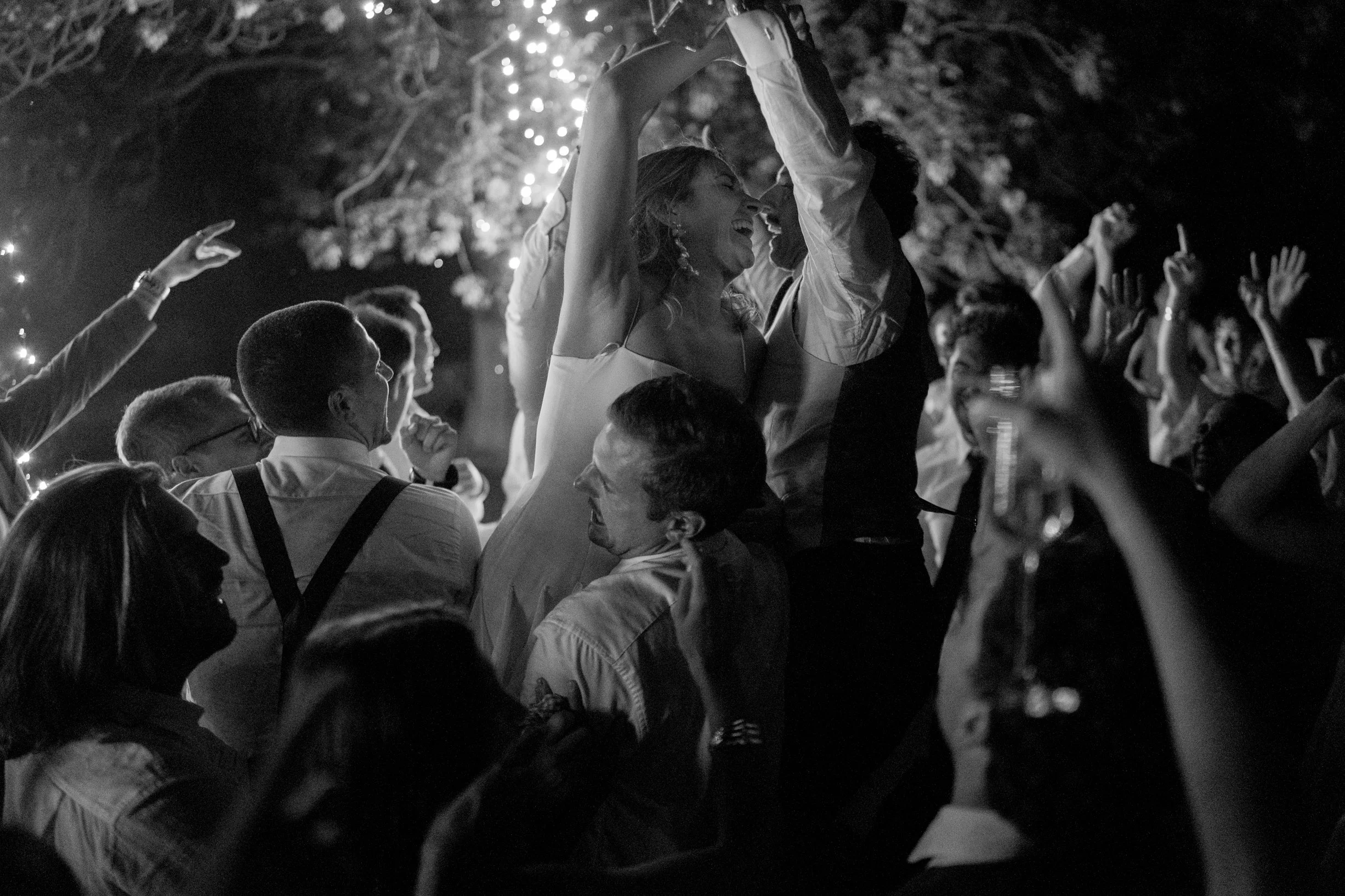 A black-and-white photo of a lively outdoor celebration with people dancing and smiling under string lights, with a woman and a man in the center sharing a joyful moment.