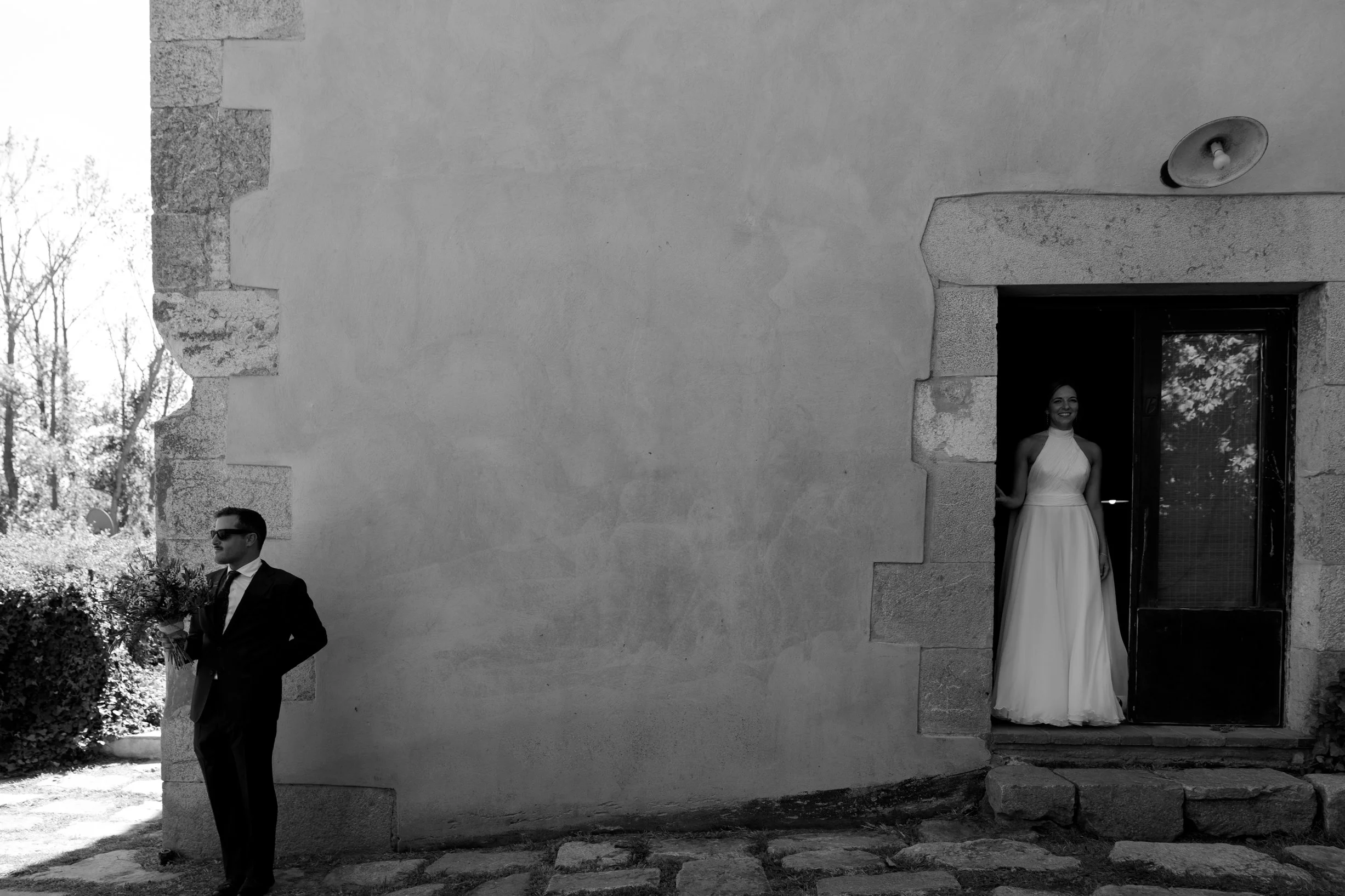 A black and white photo of a man in a suit with sunglasses holding a bouquet of flowers, standing outdoors near a stone wall, with a woman in a wedding dress standing in a doorway behind him, smiling.