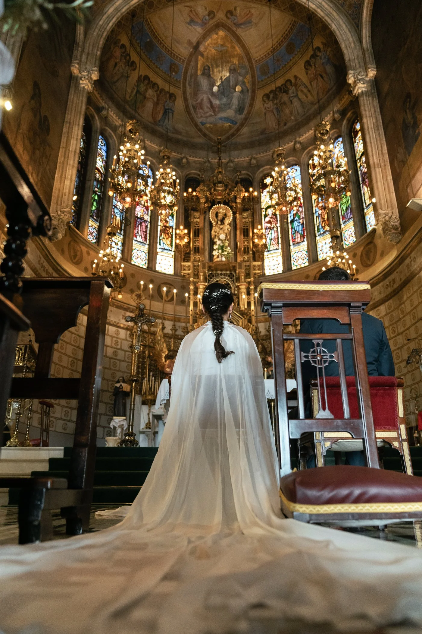 A bride in a white wedding gown standing at the altar in a church with ornate stained glass windows and gold chandeliers, seen from behind.