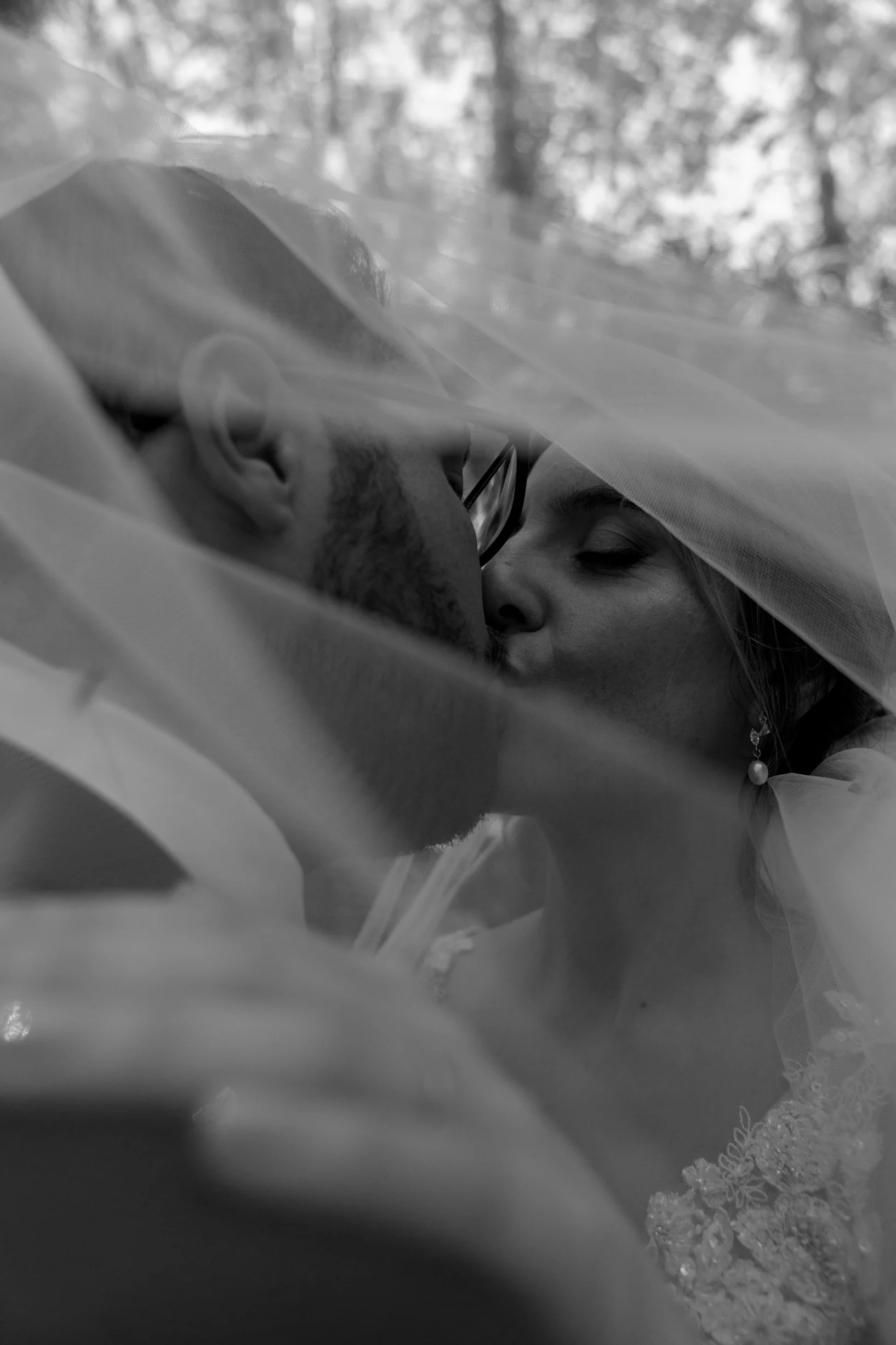 A black and white photo of a couple kissing, viewed through a sheer fabric, outdoors with trees in the background. The man has glasses and a beard, and the woman has earrings and makeup, wearing a wedding dress.