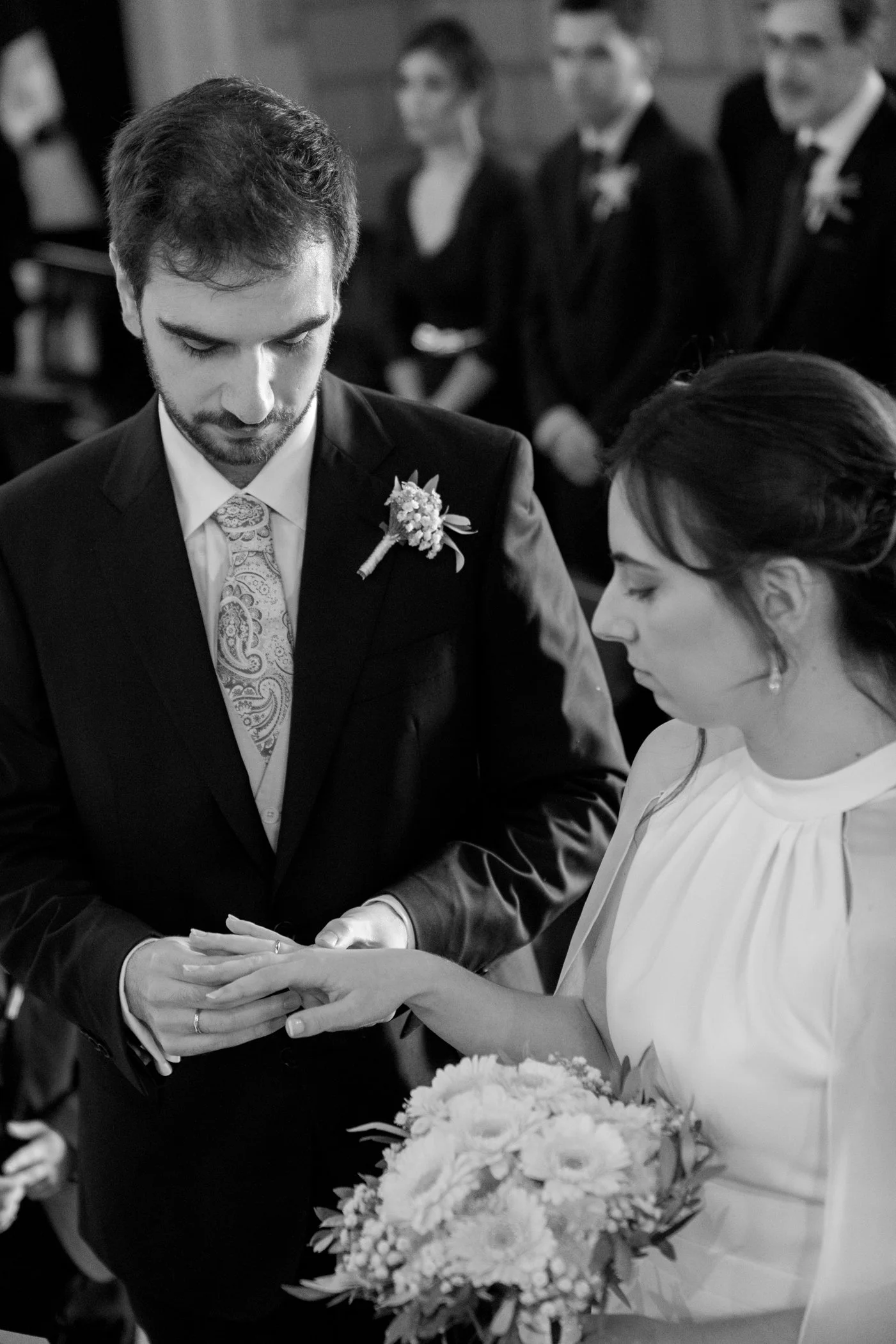 A black-and-white photo of a couple during their wedding ceremony, with the groom placing a ring on the bride's finger. The groom wears a dark suit with a boutonniere, and the bride wears a white dress and holds a bouquet of flowers. In the backgroun