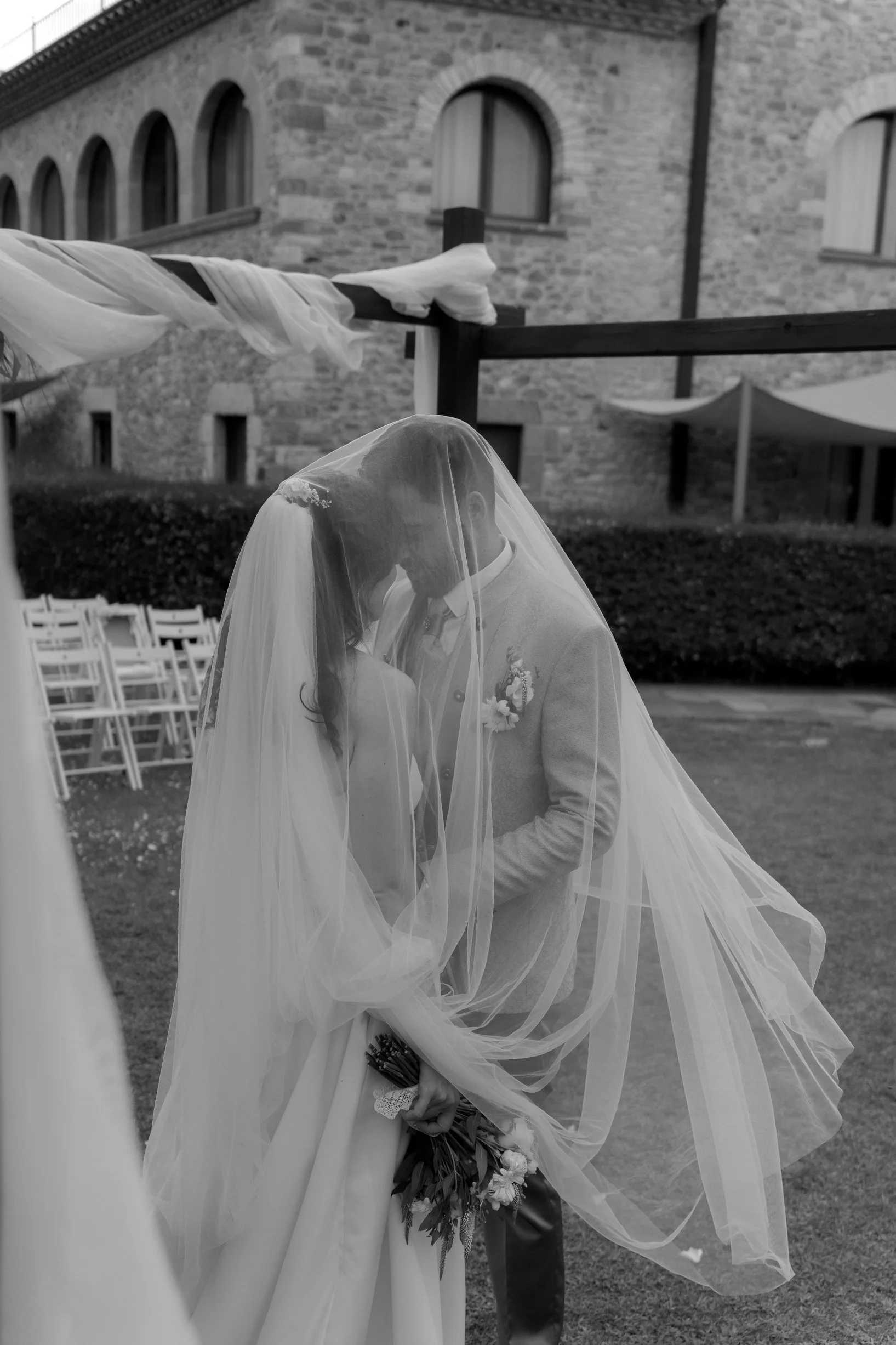 A bride and groom sharing a kiss and holding a bouquet during their wedding ceremony outdoors, with a building and chairs in the background.