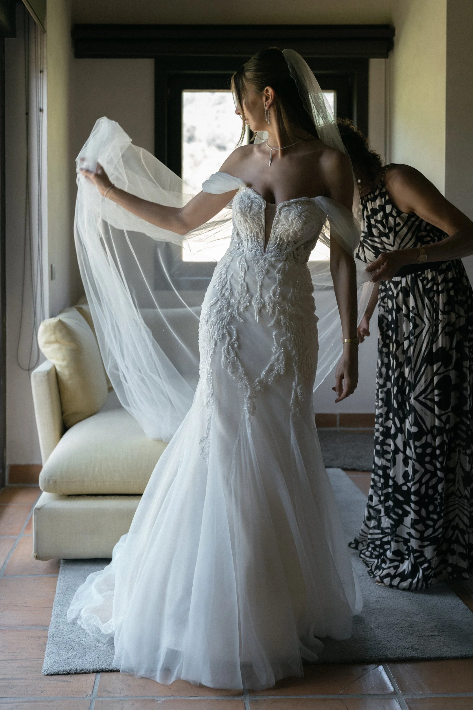 A bride in a white wedding dress is being assisted by a woman in a patterned dress. The bride is holding the end of her veil, and sunlight is coming through a window behind her.