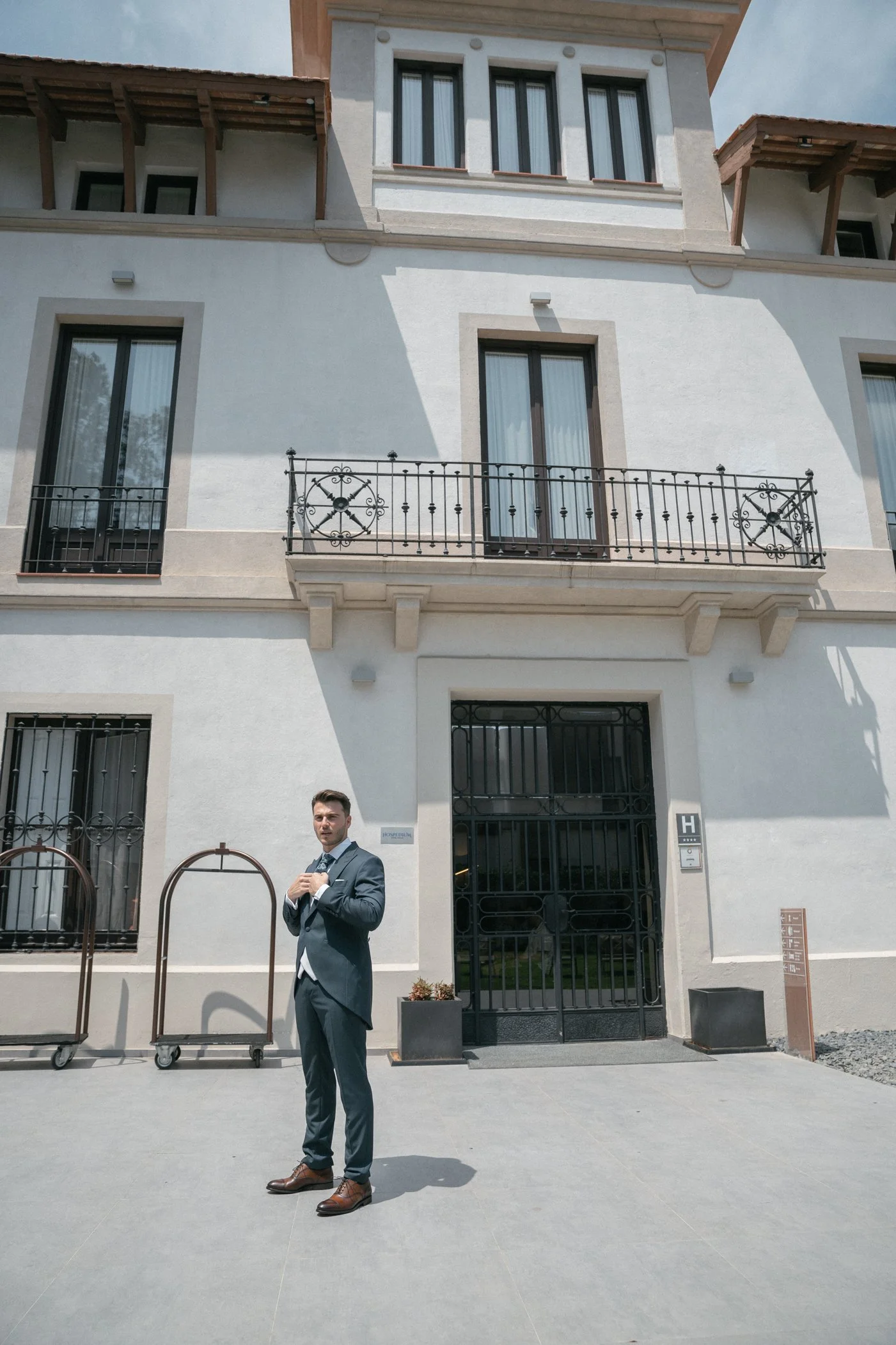 A man in a suit stands in front of a modern white multi-story building with balconies and large windows, near a luggage cart.