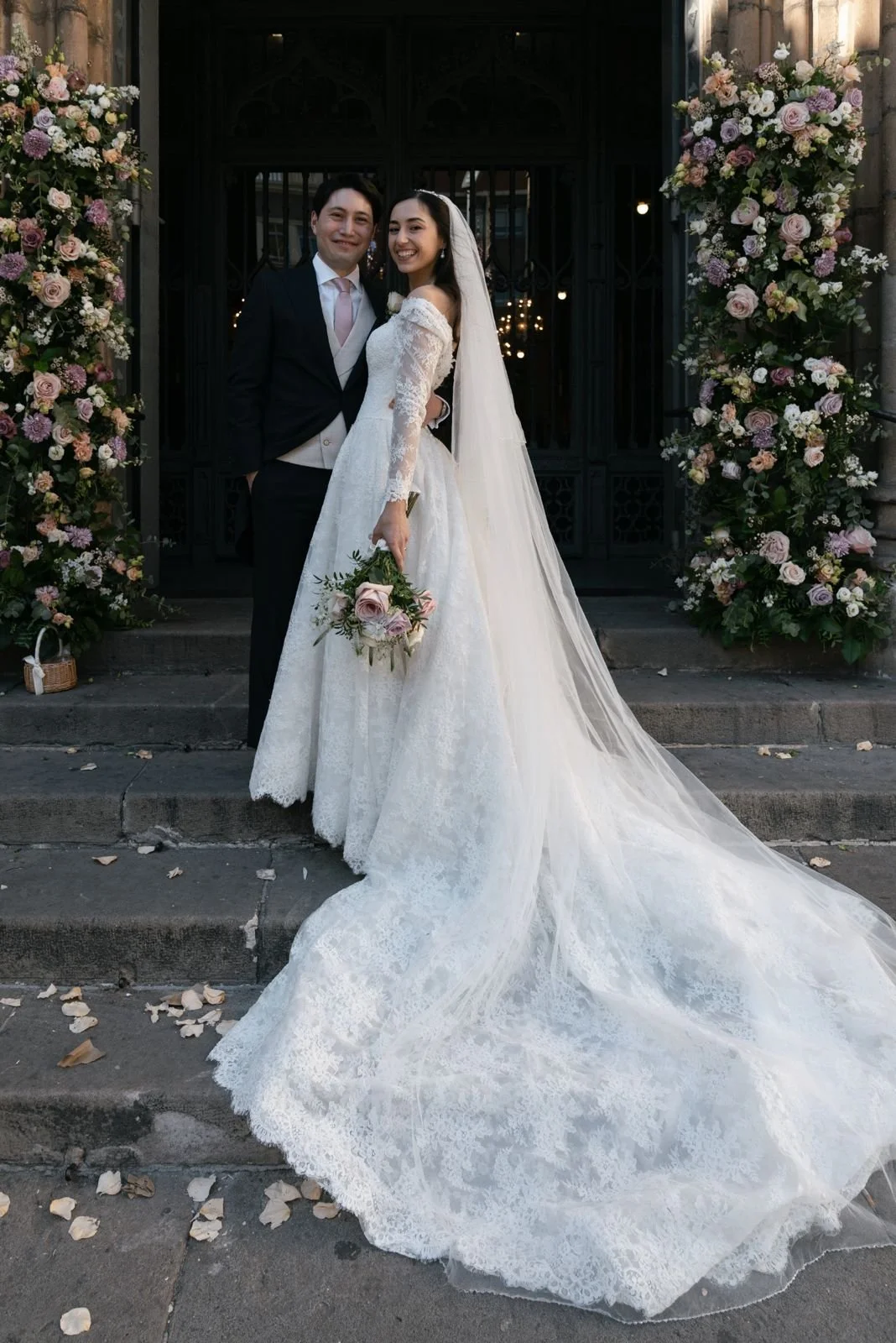 A bride and groom standing together outside a church entrance decorated with large floral arrangements. The bride is in a white lace wedding gown with a long train and veil, holding a bouquet of flowers. The groom is in a black suit with a light-colo