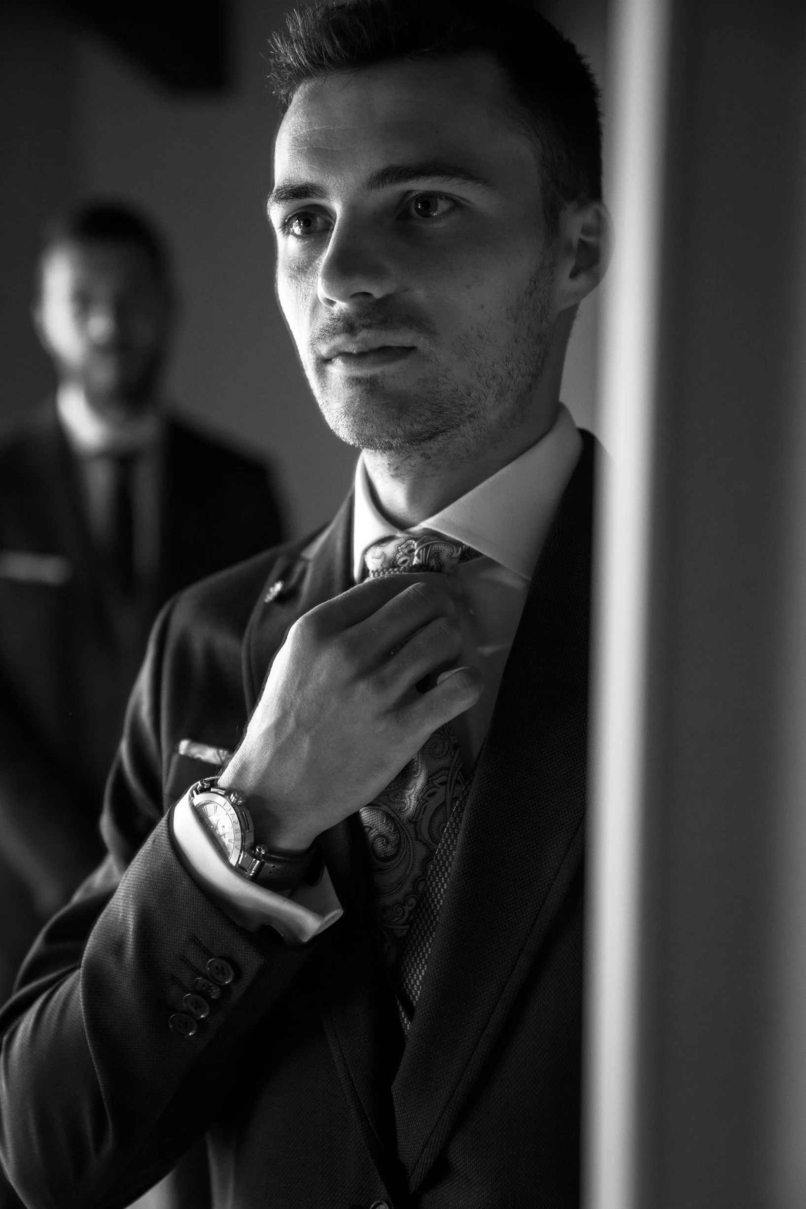 A young man in a suit adjusting his tie, standing near a wall, with another blurred man in the background.