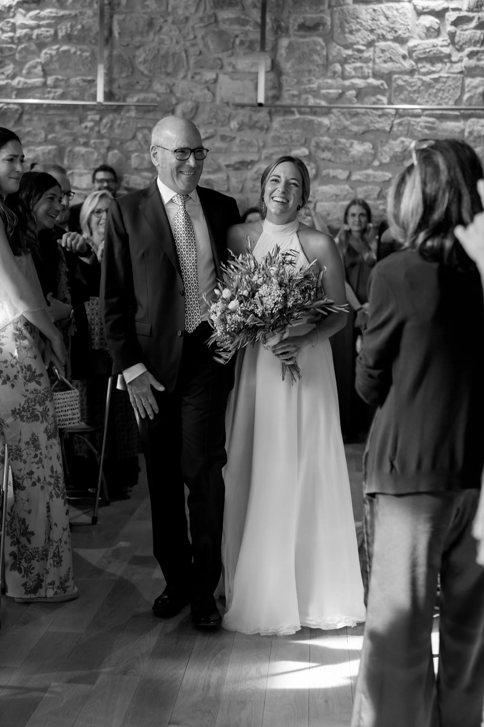 A bride holding a bouquet of flowers walking down the aisle with a man, likely her father, in a suit, at her wedding with guests smiling in the background.
